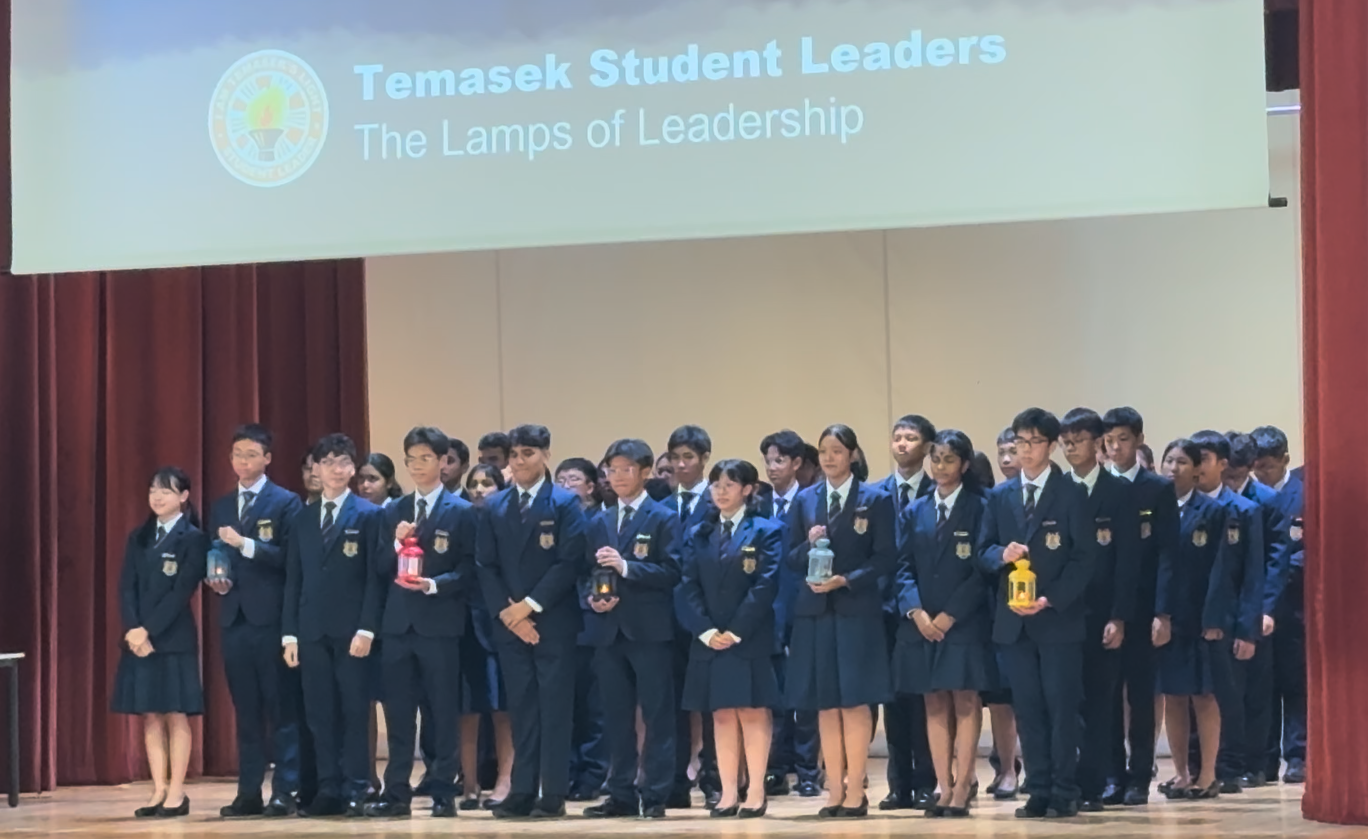 Students in navy uniforms stand on stage holding lanterns under a screen reading Temasek Student Leaders, The Lamps of Leadership.