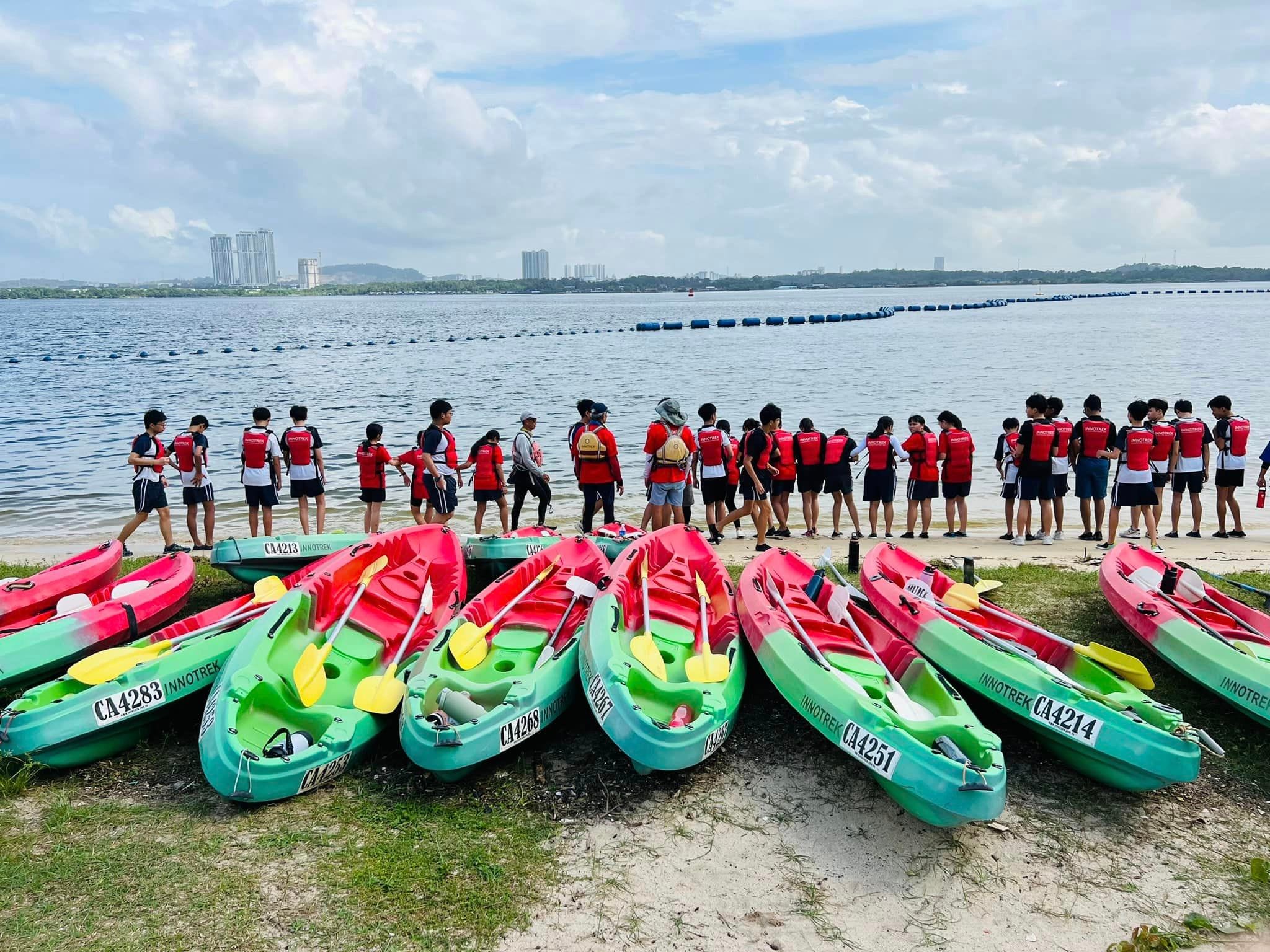 A group of students in red life vests stand near colorful kayaks on a sandy shore beside a wide calm waterway.