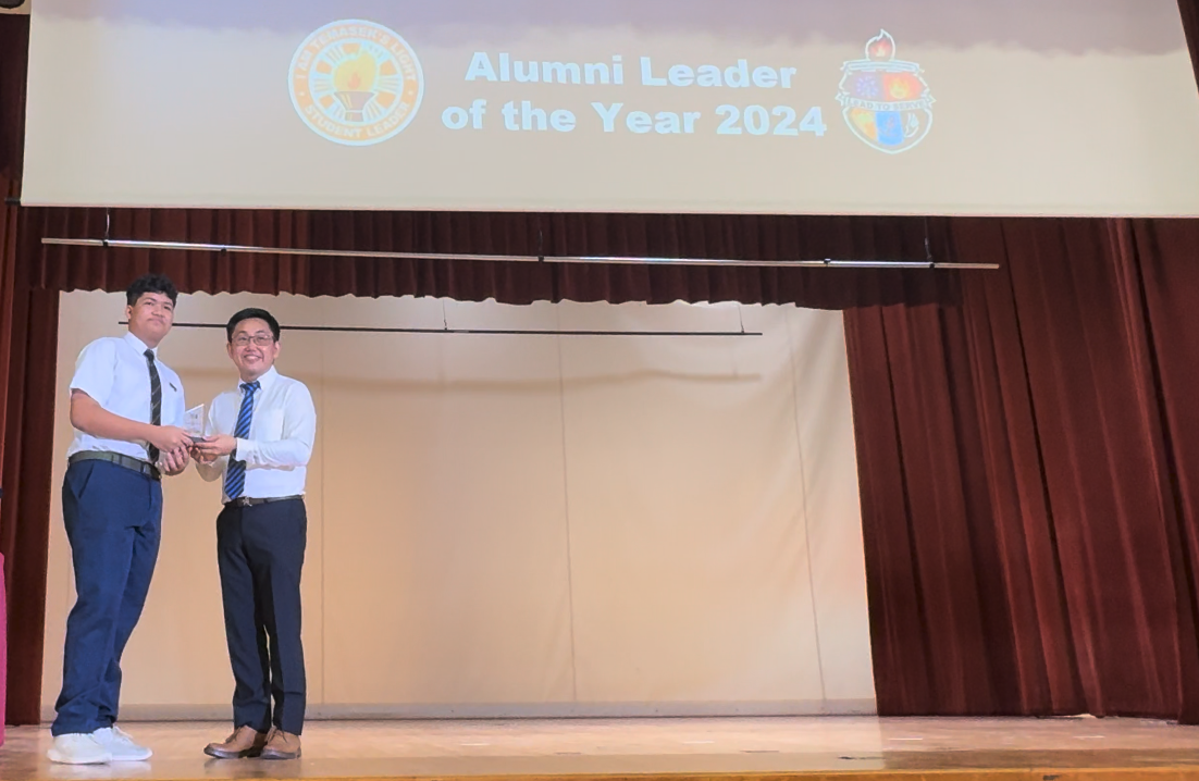 Two men in white shirts and ties stand on a stage holding a trophy under an Alumni Leader of the Year 2024 banner.
