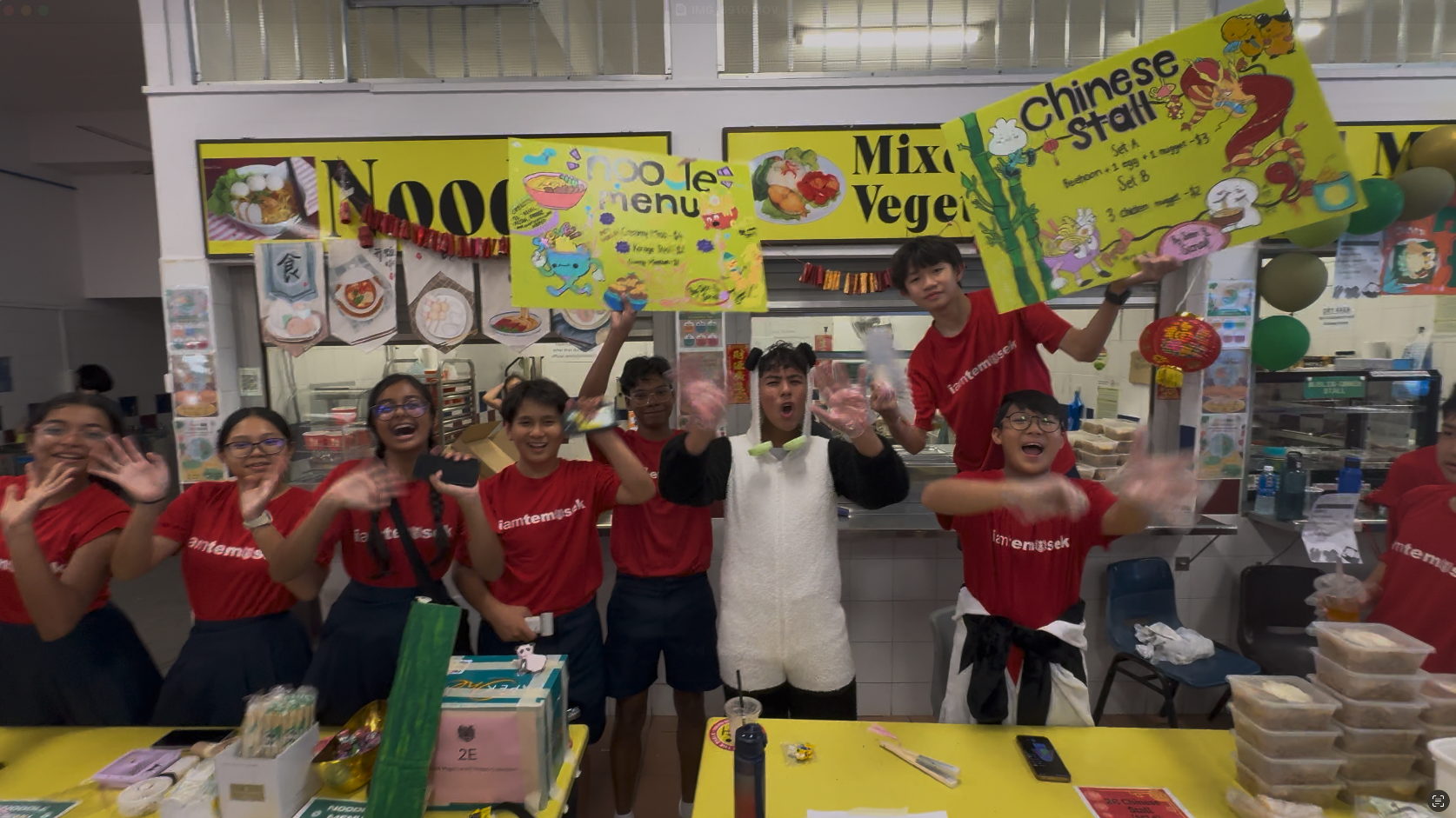 Students in red Temasek shirts cheer at a school food fair with colorful Chinese stall and noodle menu banners.