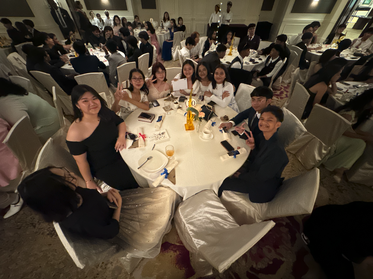 A group of formally dressed young people smiling at a round dinner table in a large banquet hall.