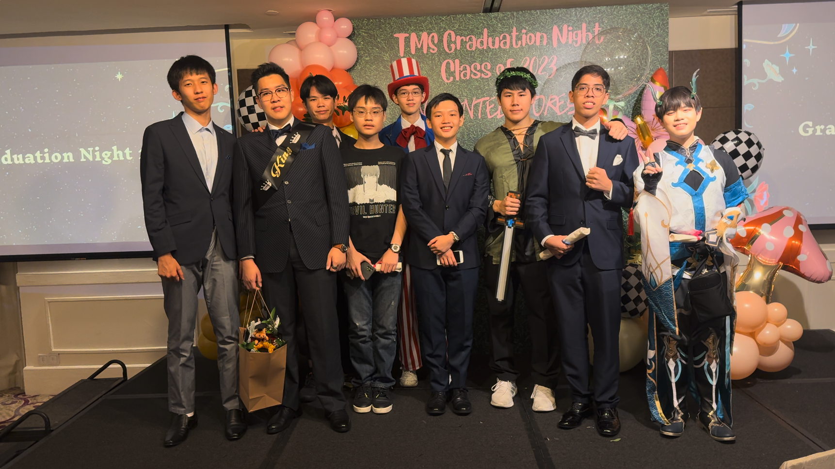 Eight young men pose at TMS Graduation Night Class of 2023, some in suits and one in costume, with balloons behind them.