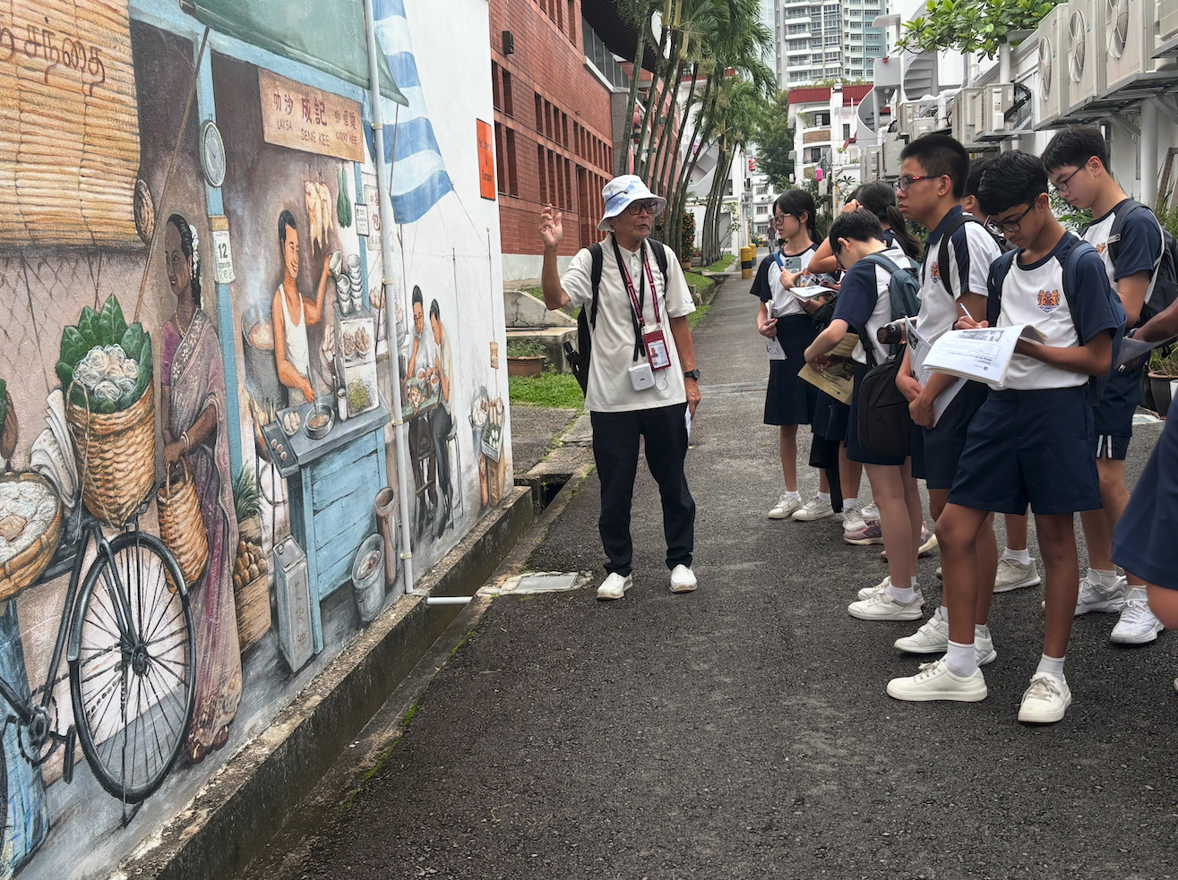 A guide in a white hat explains a colorful street mural depicting market scenes to a group of students taking notes.