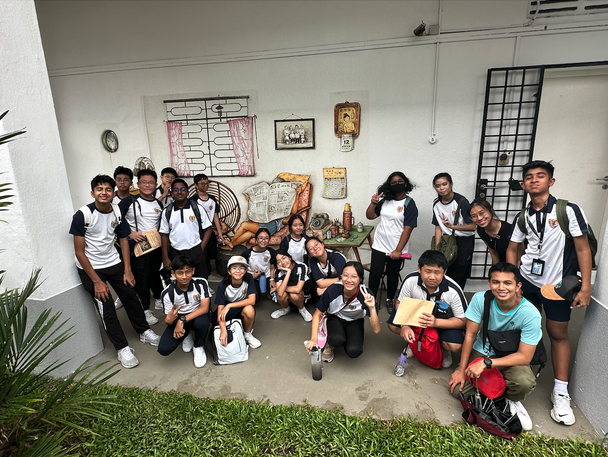 A group of teenagers in matching white and navy uniforms pose in front of a detailed wall mural of a vintage room scene.