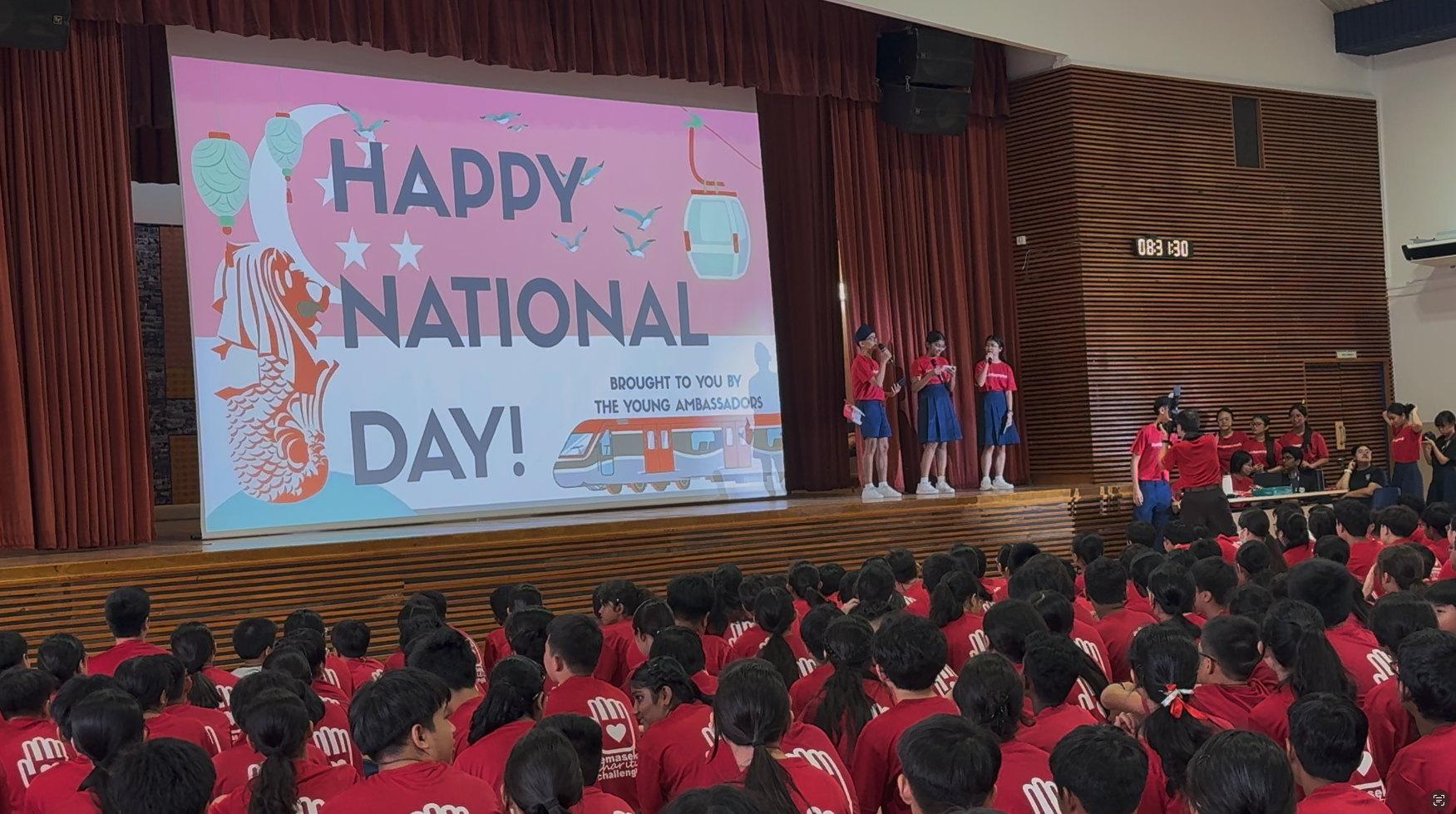 Students in red shirts sit in a hall facing a stage with a Happy National Day projection and student presenters.