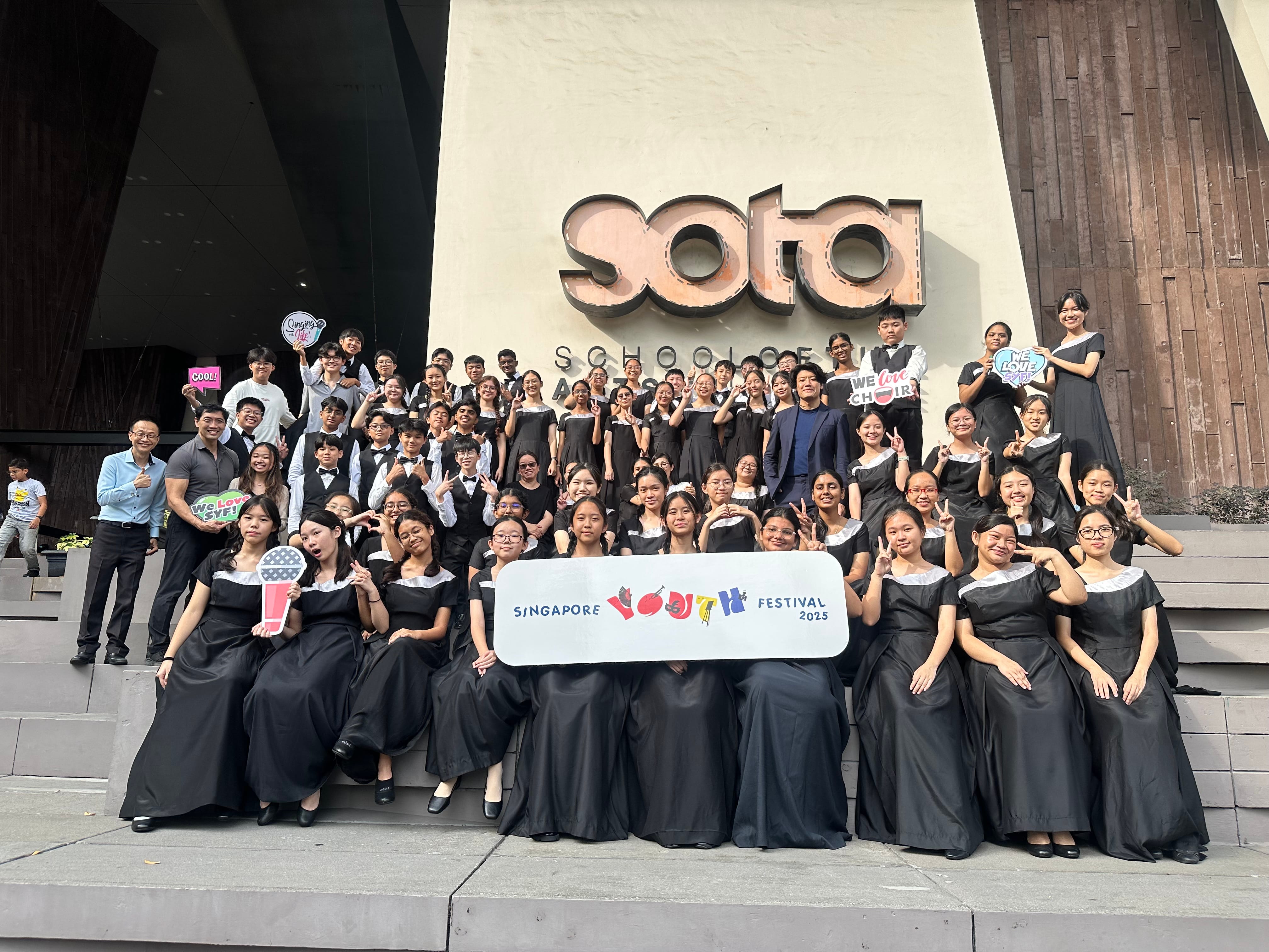 Large choir group in black uniforms posing on steps at SOTA building, holding a Singapore Youth Festival 2025 banner.