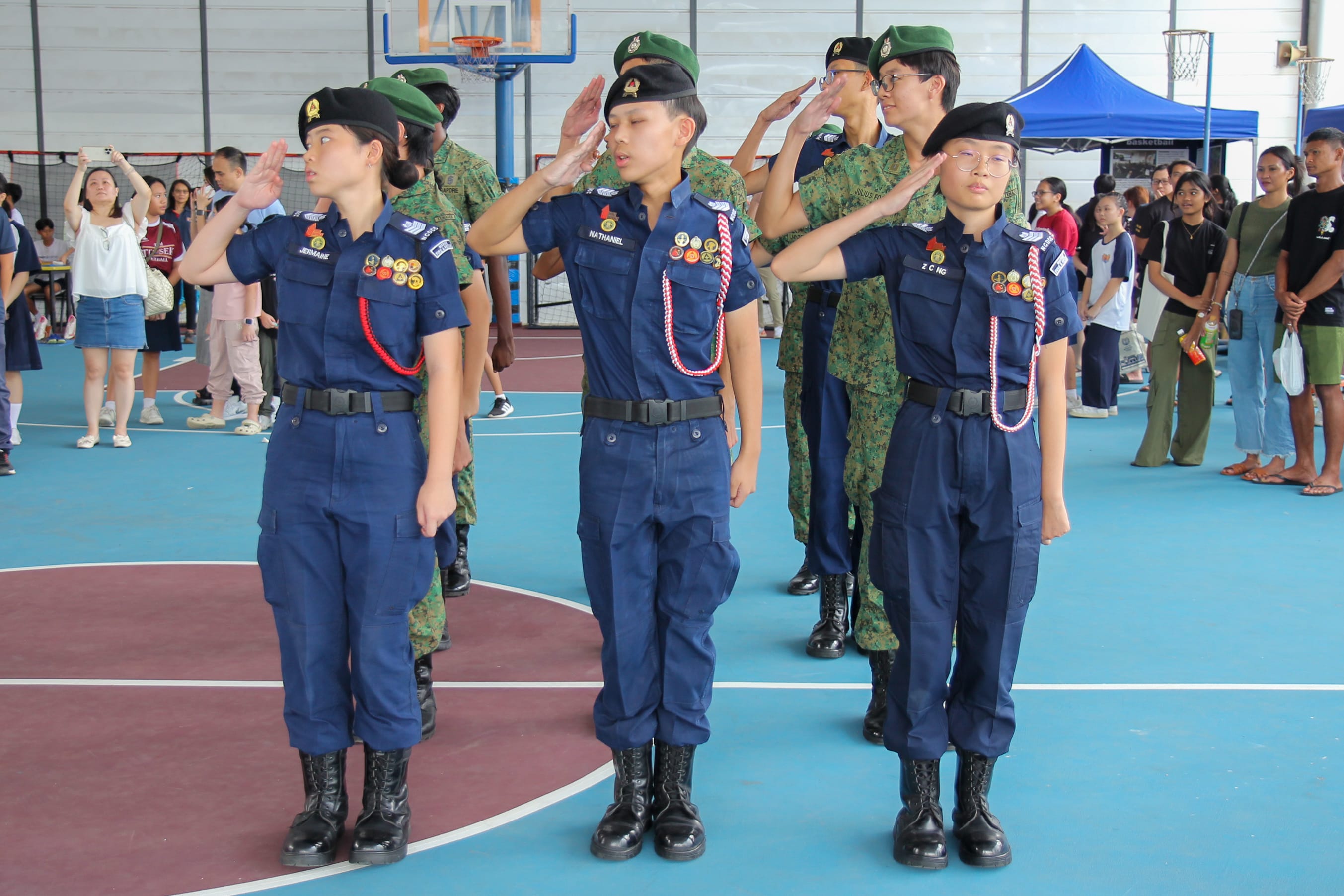 Young cadets in blue uniforms and berets saluting during a ceremony on an indoor basketball court with spectators watching.