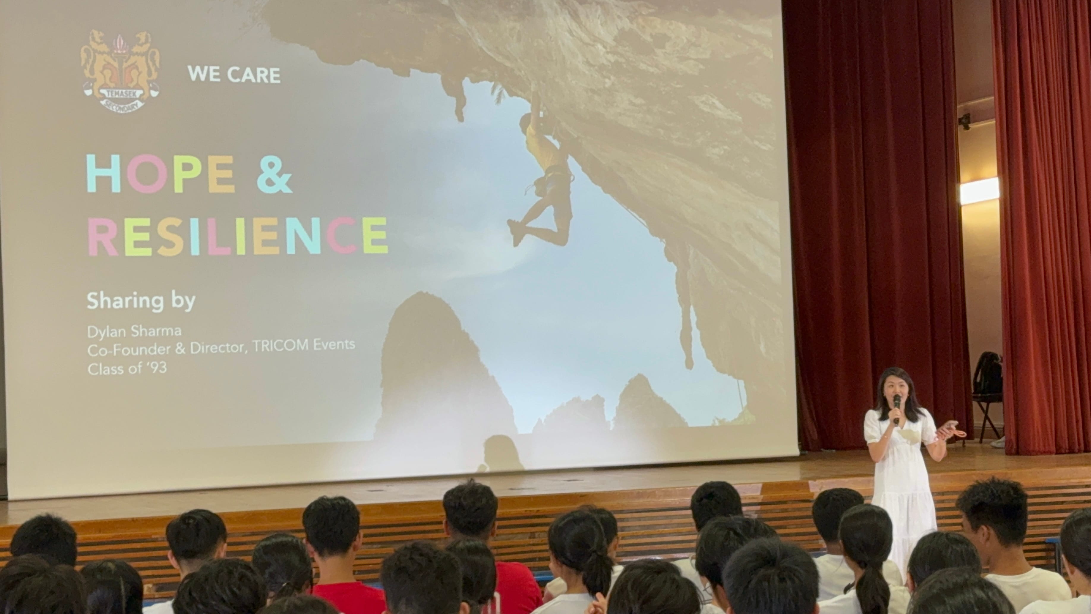 A woman in white speaks on stage before students, with a projected slide reading "Hope & Resilience" behind her.