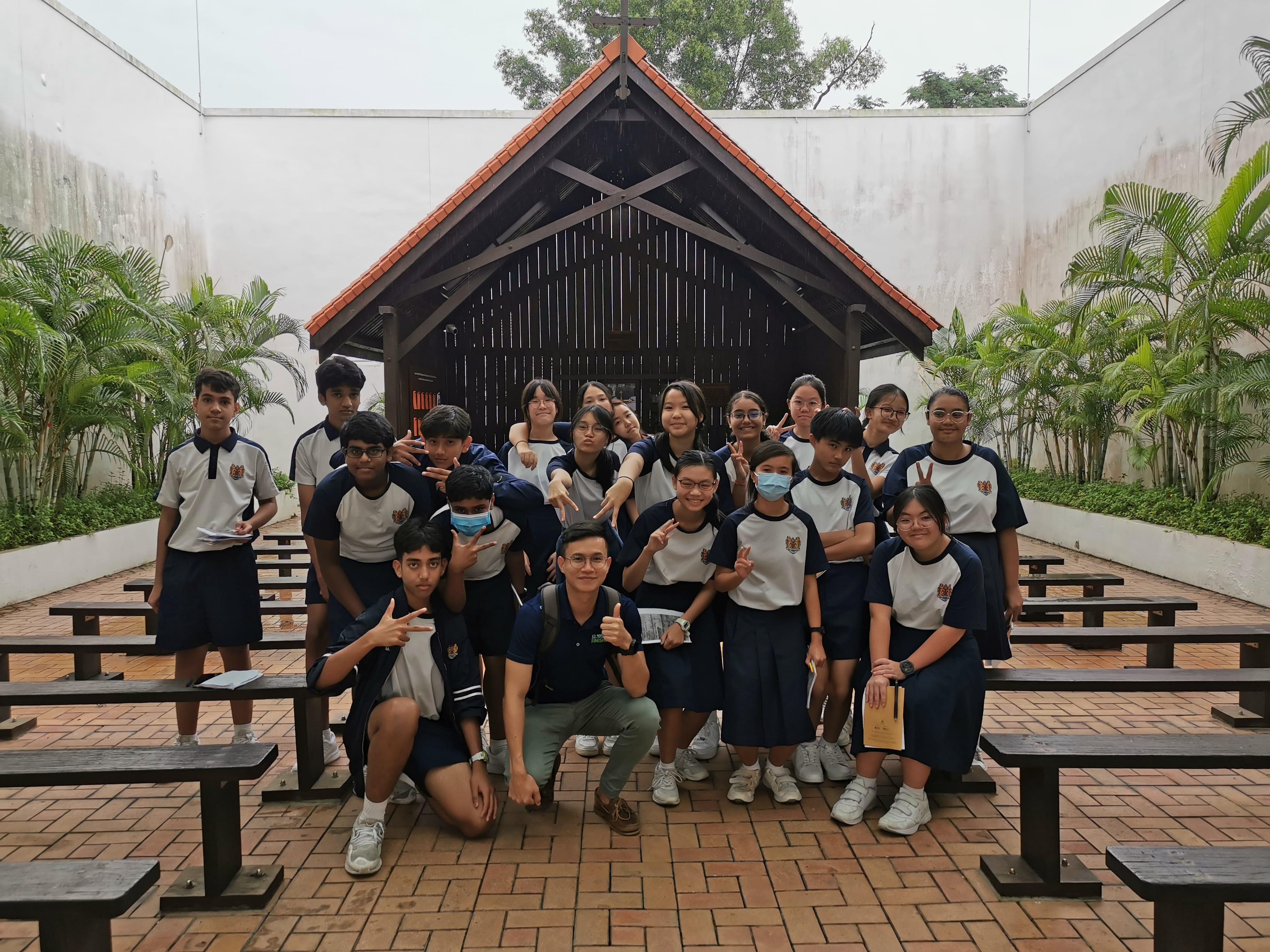 A group of students in navy and white uniforms posing together in a courtyard with a wooden chapel structure behind them.