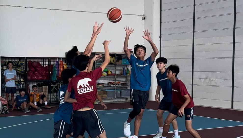 Several teenage boys play basketball indoors, with one player in blue jumping to shoot while others reach up to block.