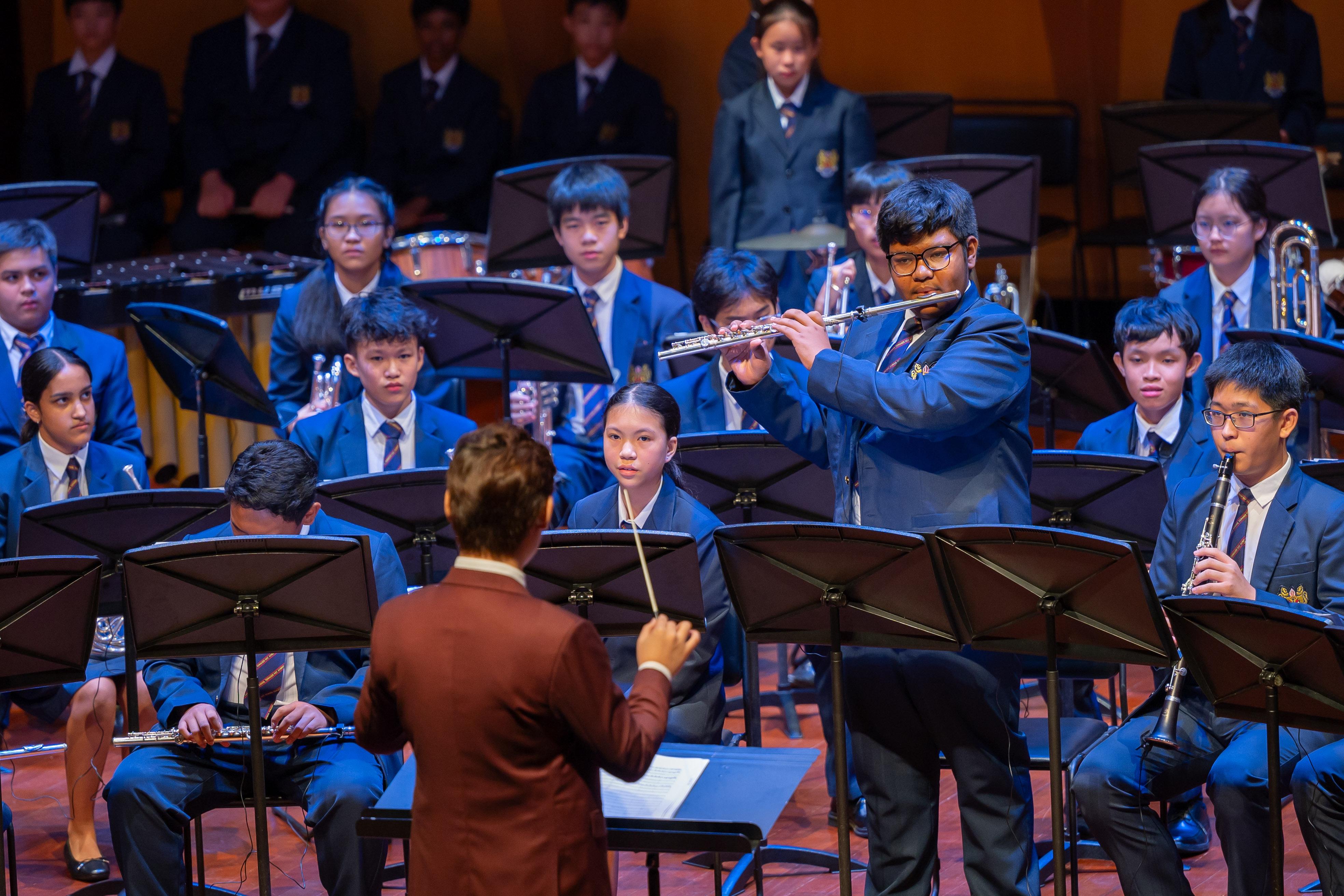 A conductor leads a school student band in blue uniforms playing flutes, clarinets, and brass instruments on stage.