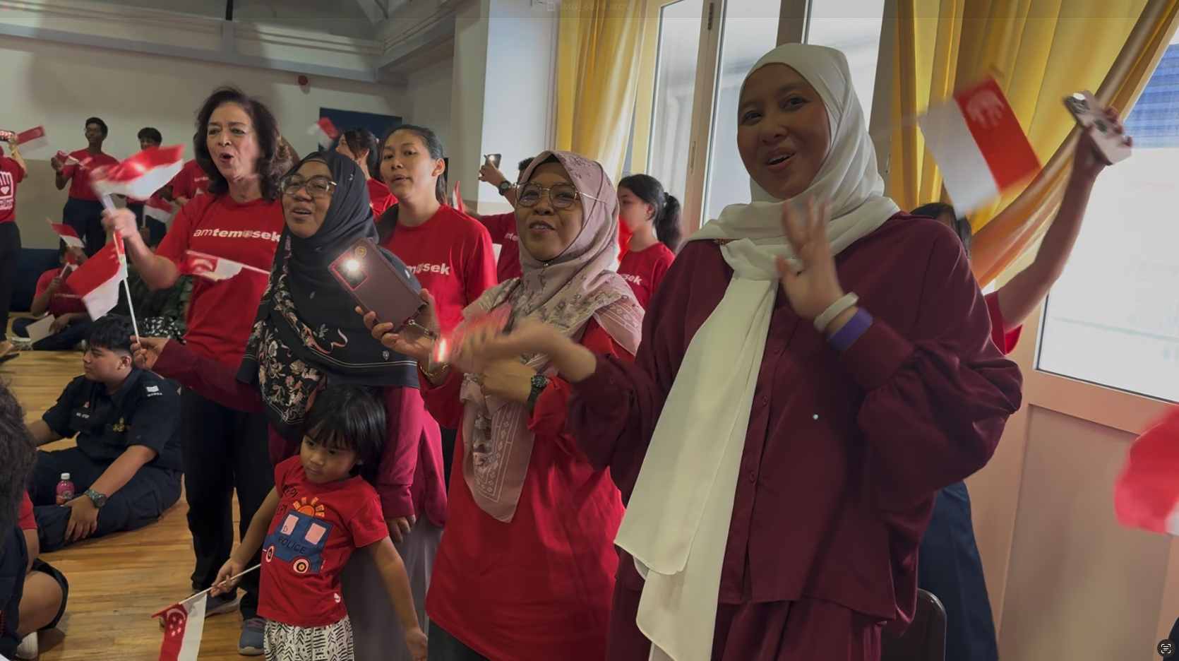A crowd of people in red shirts wave small Singapore flags and cheer indoors at a festive gathering.