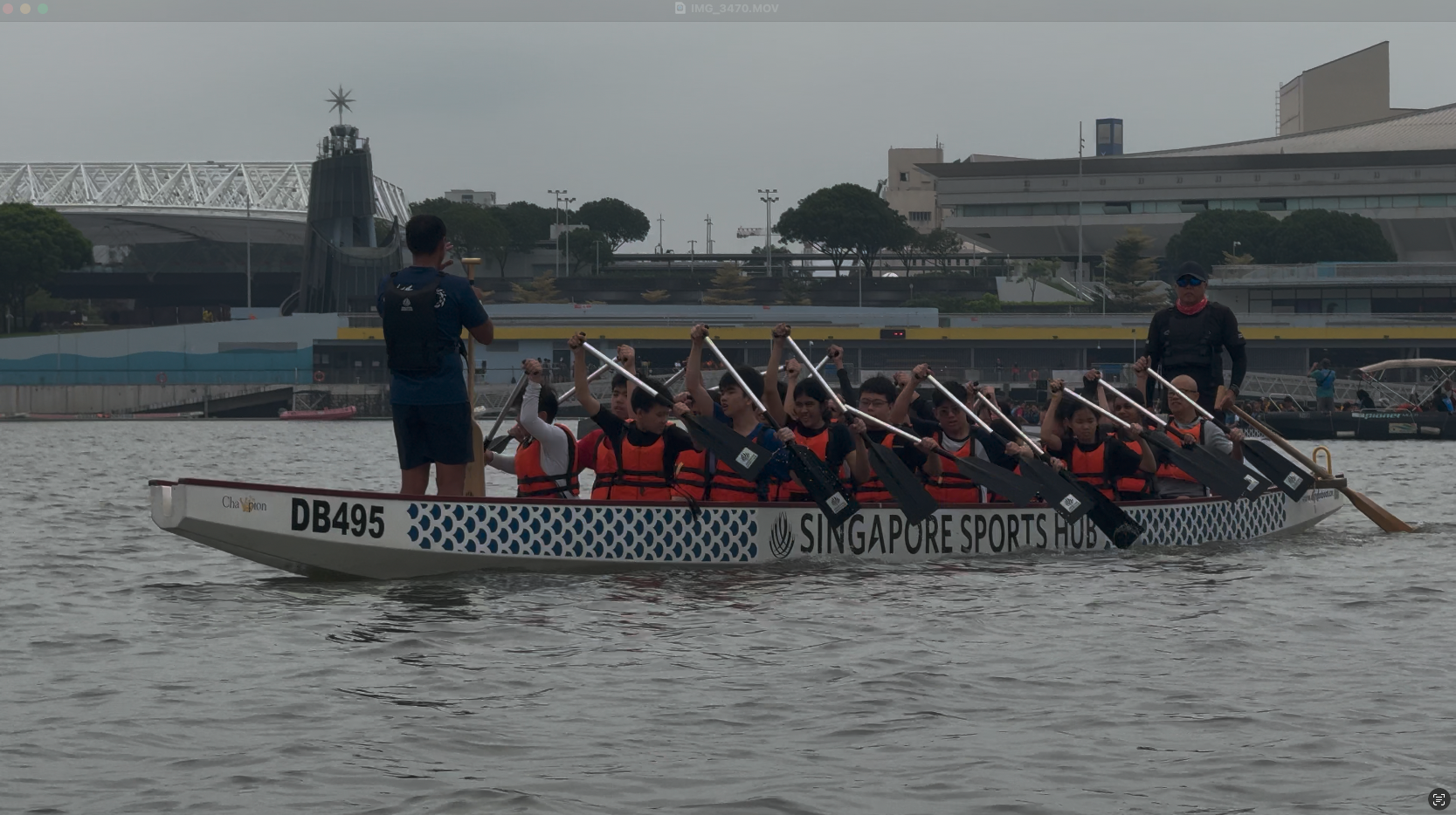 A team in orange life vests paddles a dragon boat labeled DB495 Singapore Sports Hub on open water.