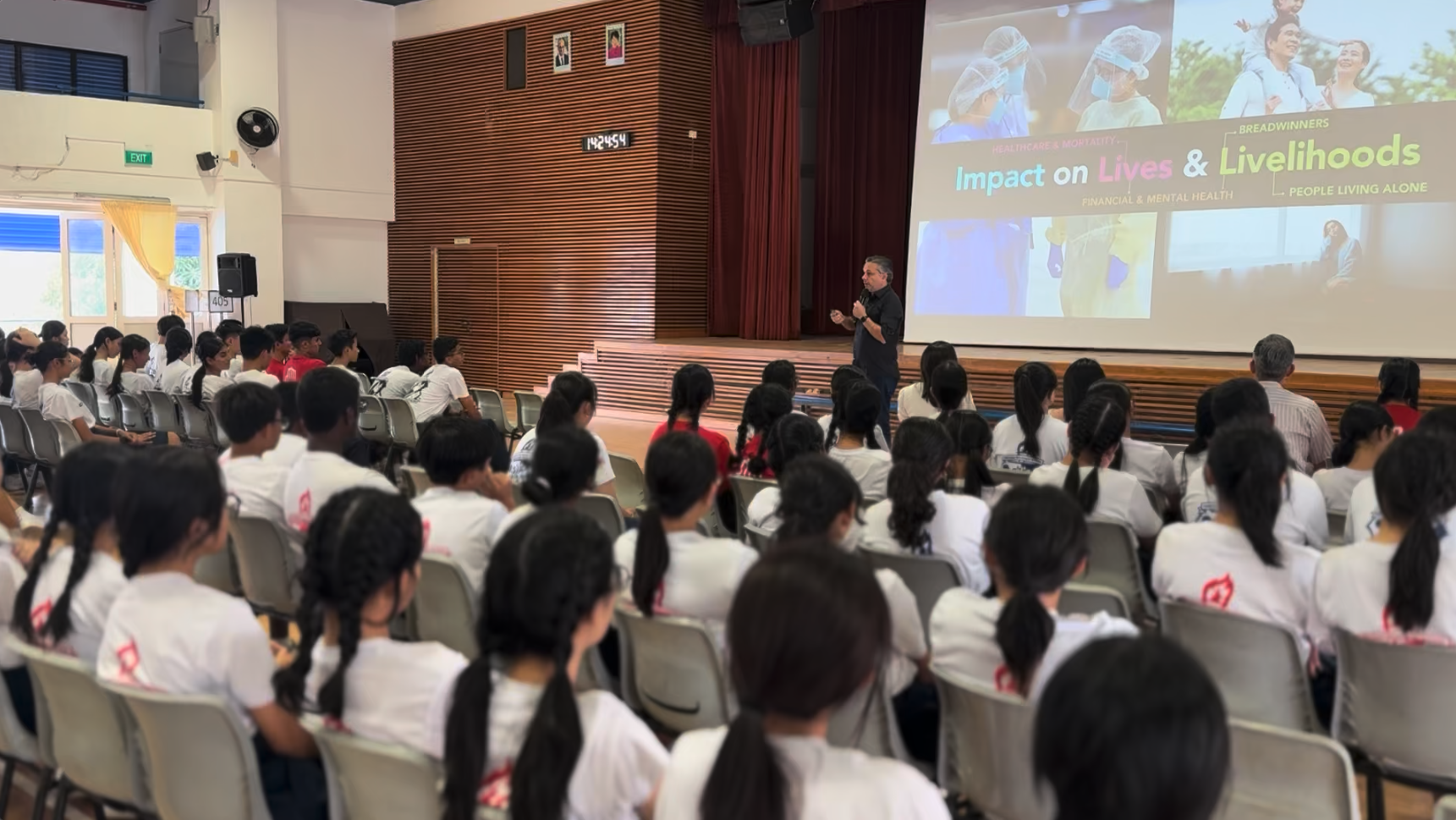 A speaker presents to students in white shirts in a hall, with a slide reading "Impact on Lives & Livelihoods."