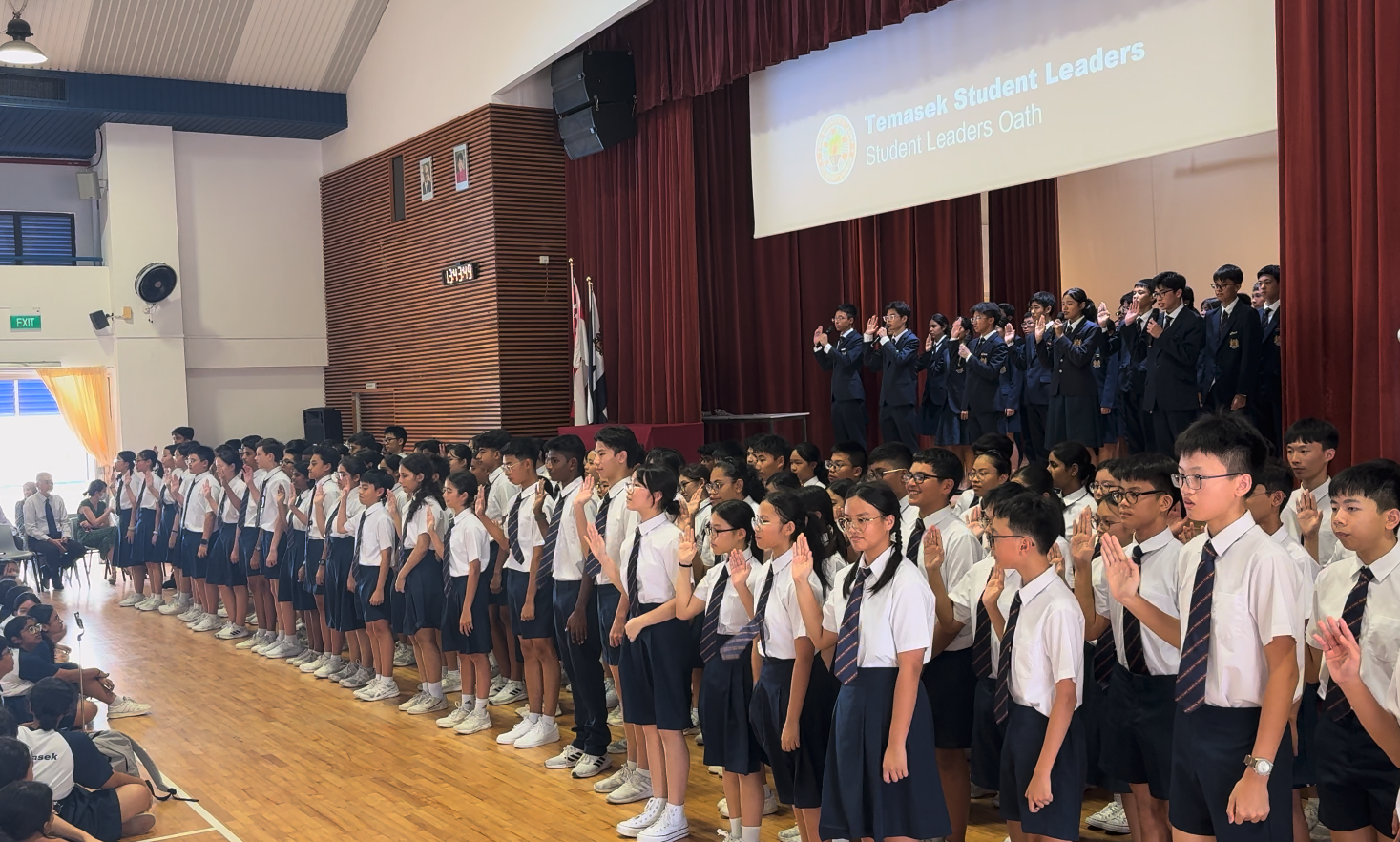 Students in school uniforms raise their right hands during the Temasek Student Leaders Oath ceremony in a hall.