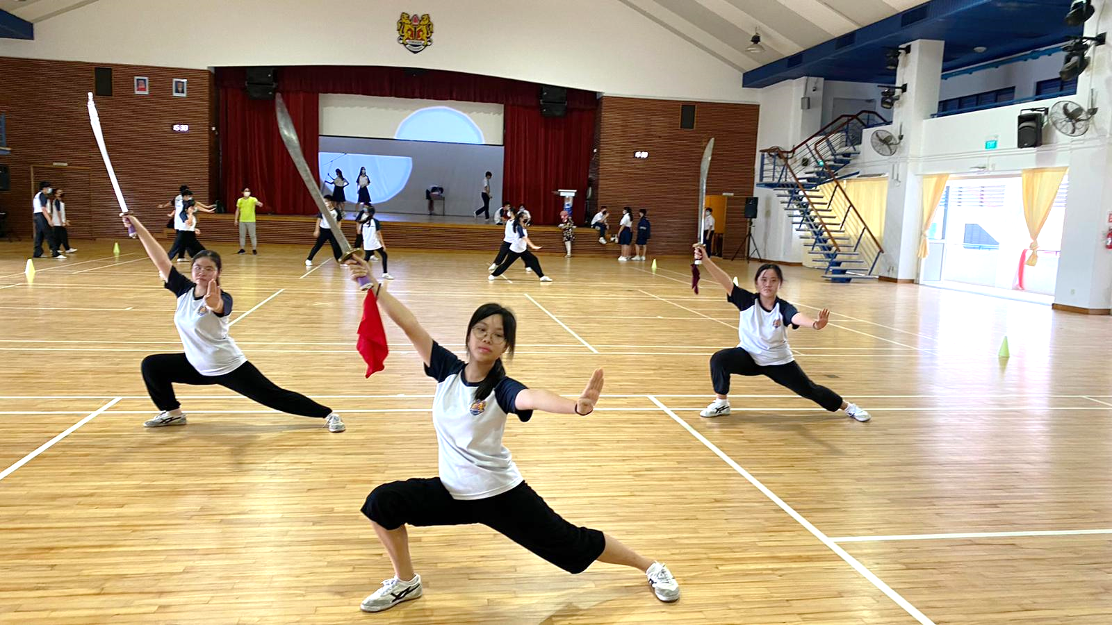 Students in white and black uniforms practice wushu sword forms in a school hall with a wooden floor.