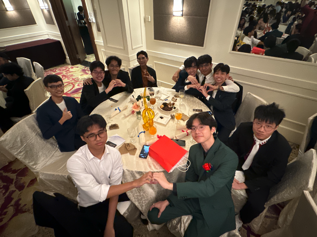 Ten young men in formal attire sit around a decorated banquet table, smiling and making hand gestures.