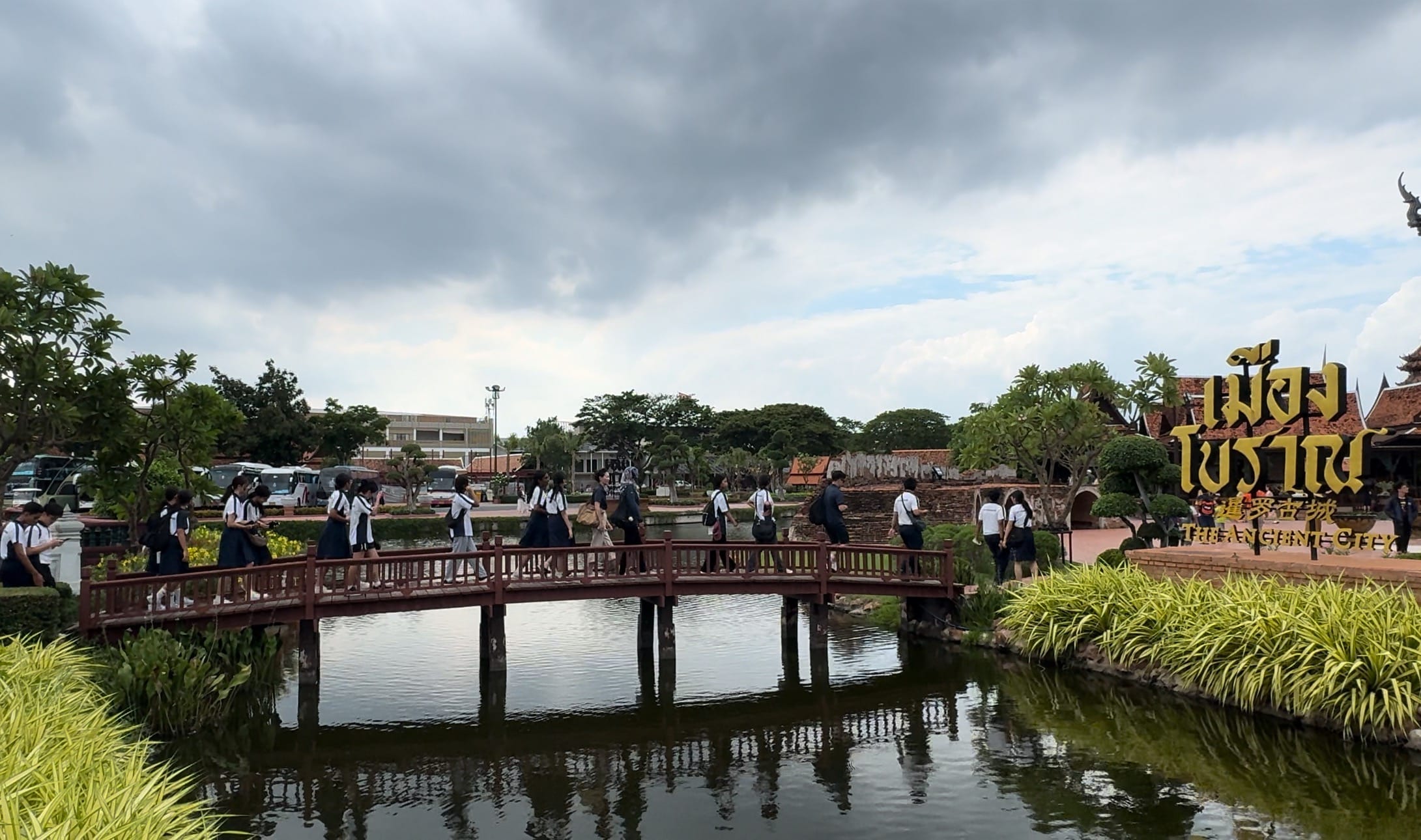 Students in uniforms walk across a wooden bridge over a pond at Ancient City Thailand under cloudy skies.
