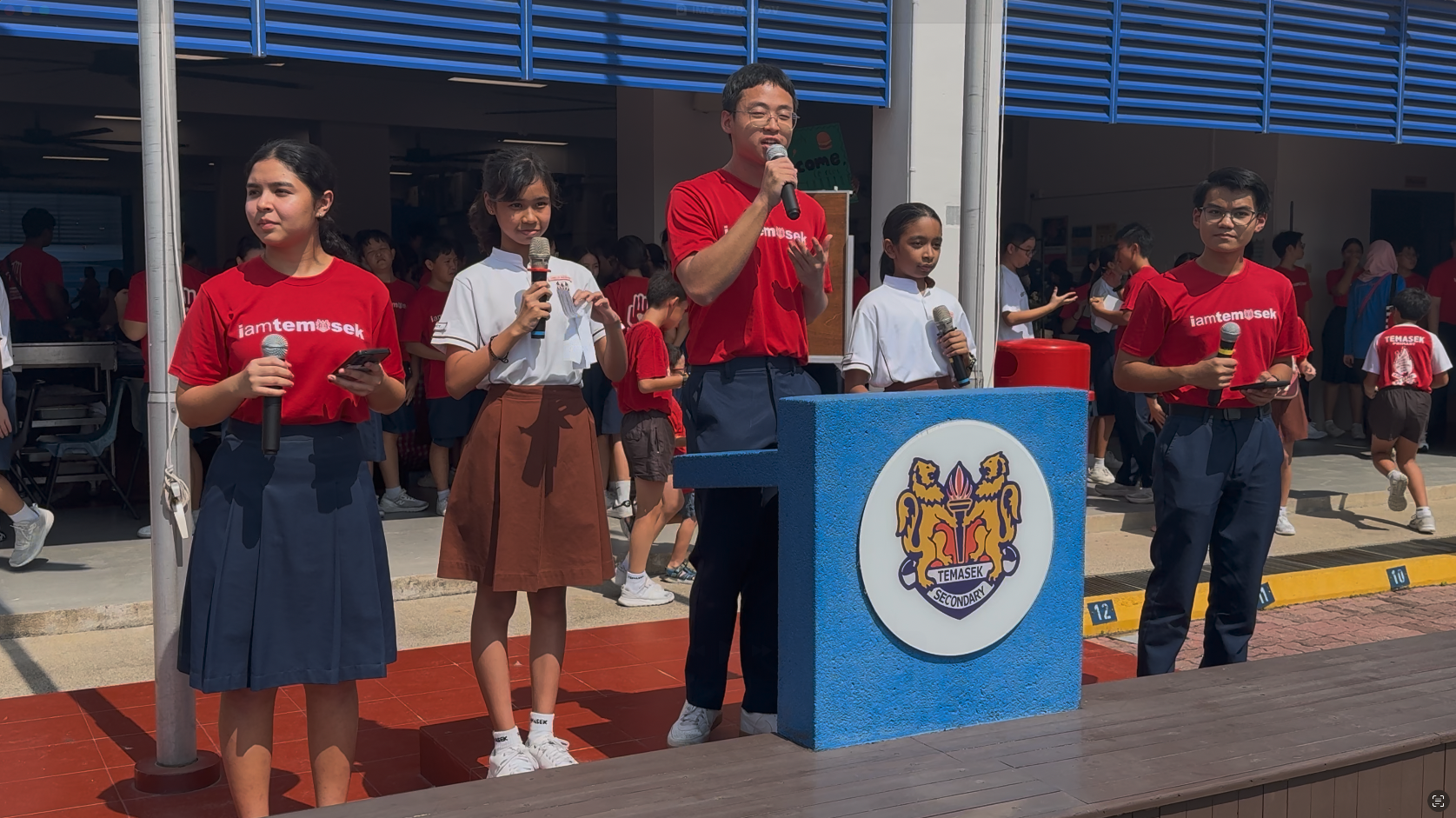 Students hold microphones on a stage at Temasek Secondary School, some in red shirts, others in uniforms.
