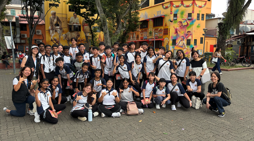 A large group of students in matching white uniforms poses outdoors in front of a colorful mural on a yellow building.