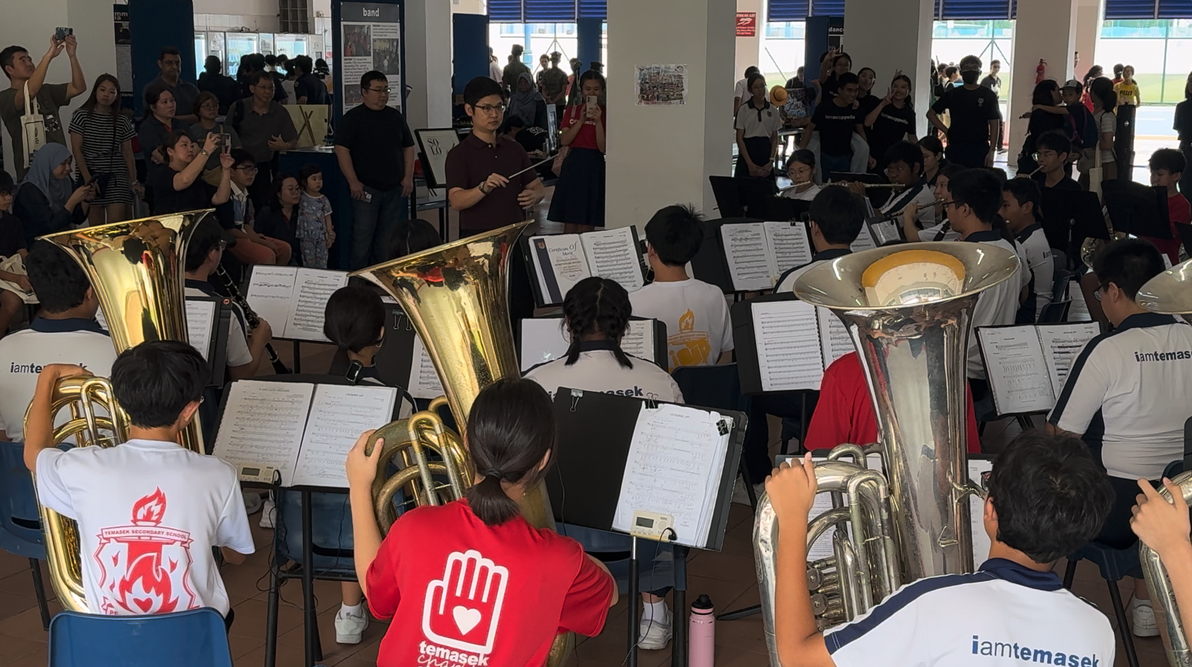 Temasek Secondary School student band playing brass instruments with sheet music, conducted by a person holding a baton.