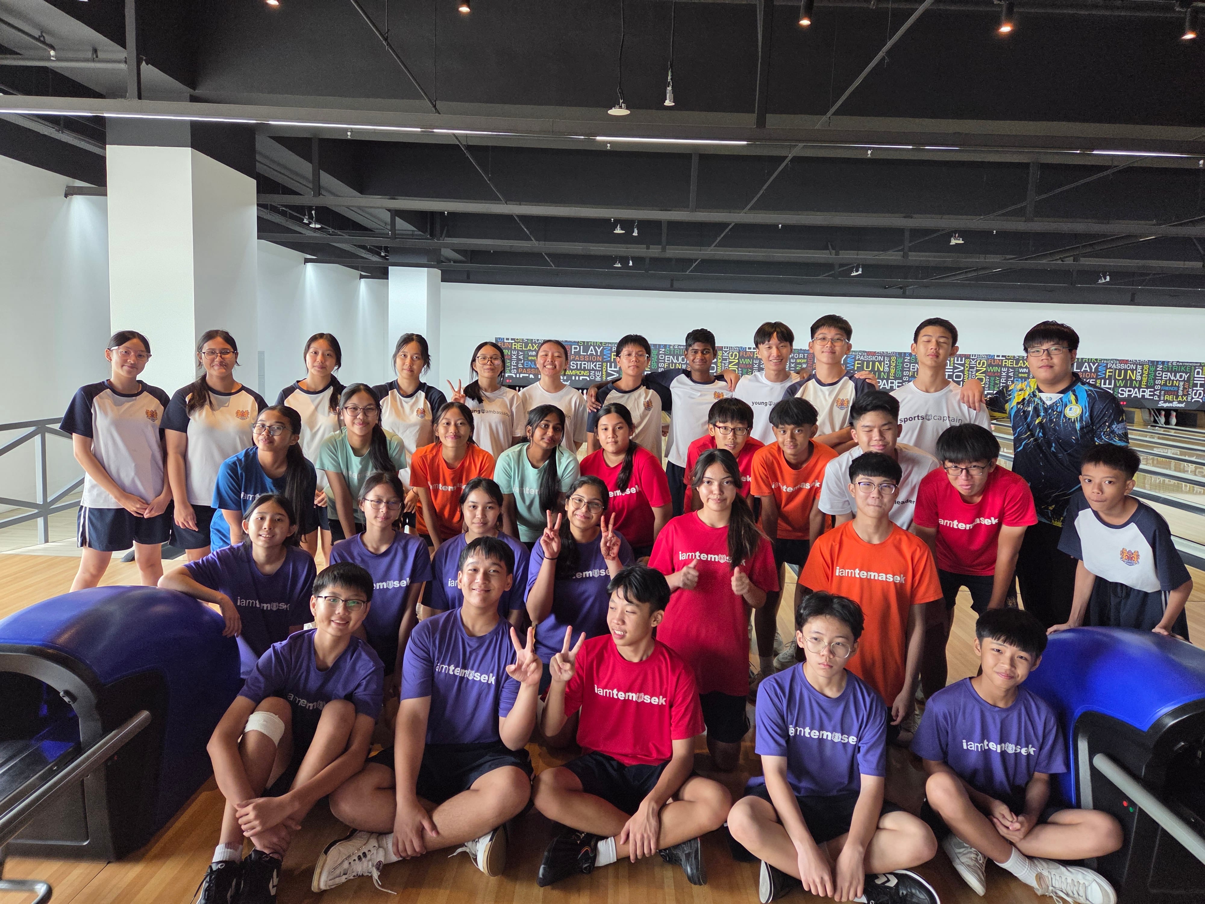 Large group of students in purple, red, and orange iamtemasek shirts posing together inside a bowling alley.