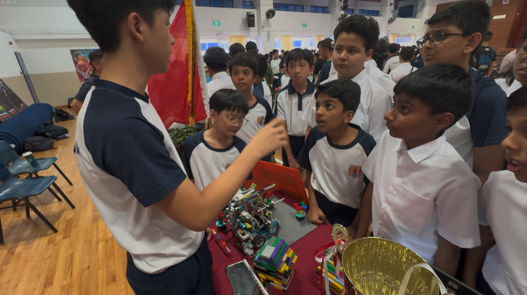 A boy in a school uniform presents a Lego robot model to a group of attentive students in a school hall.