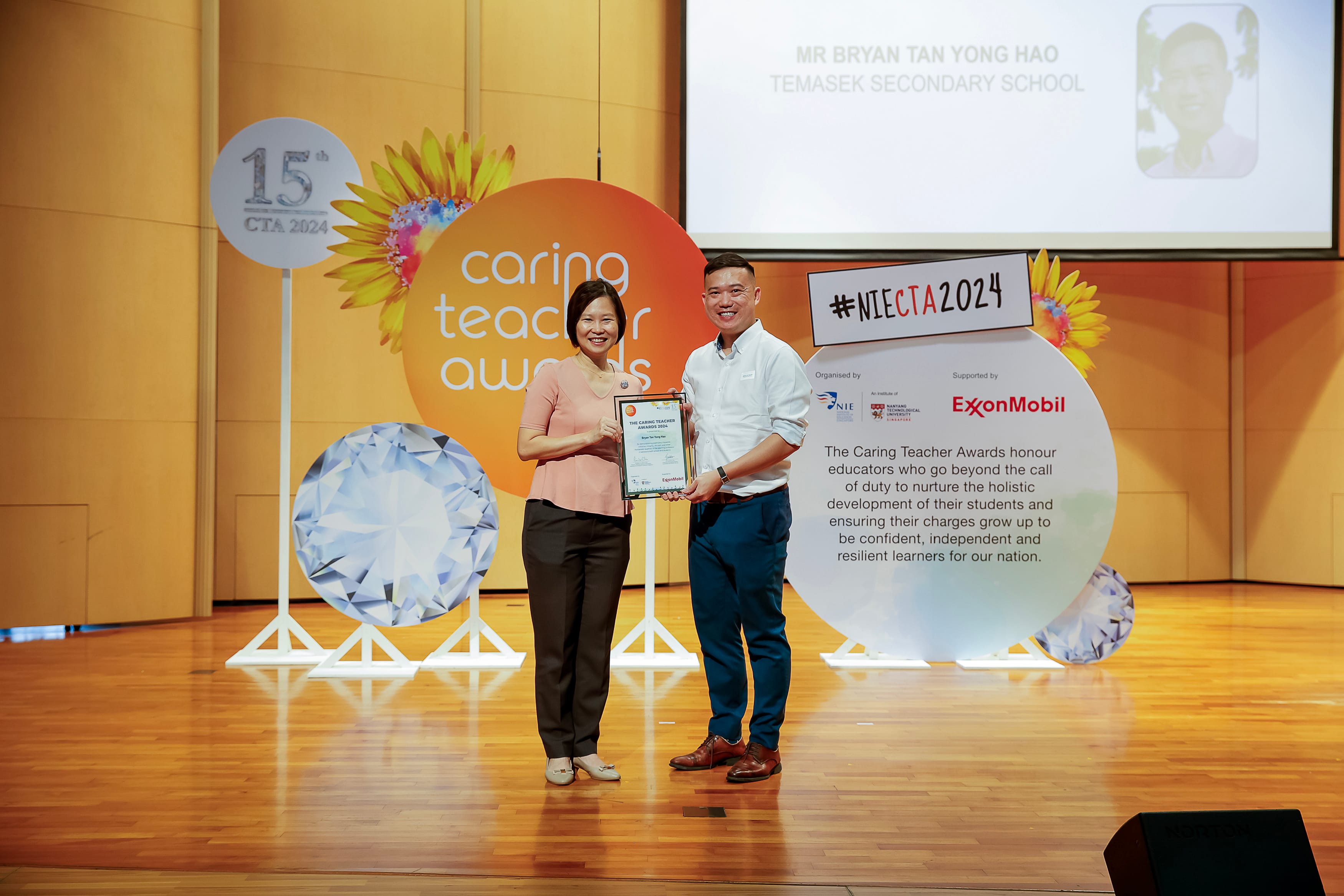 Two people on stage hold a framed certificate beside Caring Teacher Awards and #NIECTA2024 signs.