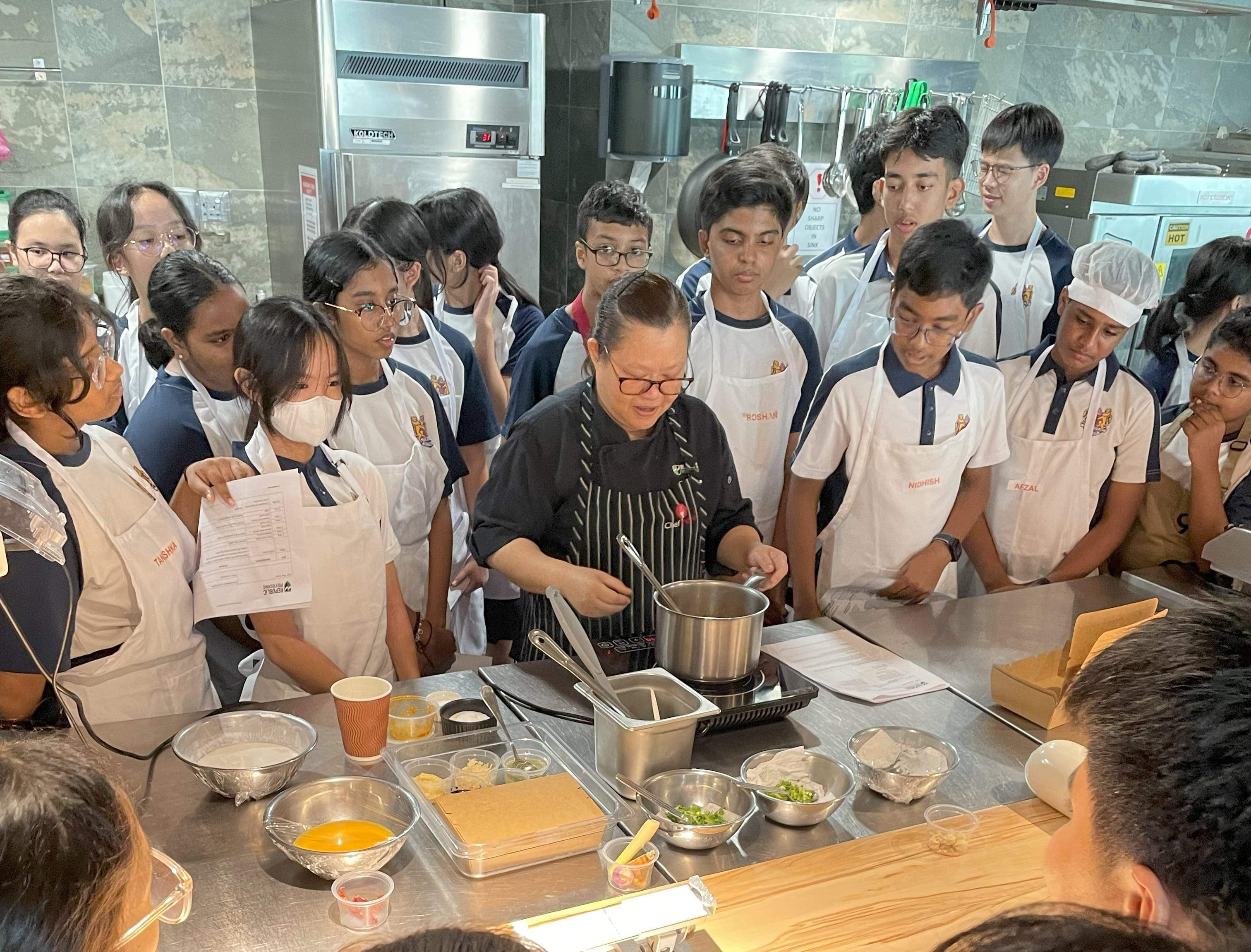 A chef in a striped apron demonstrates cooking in a pot while students in white aprons watch closely in a kitchen.