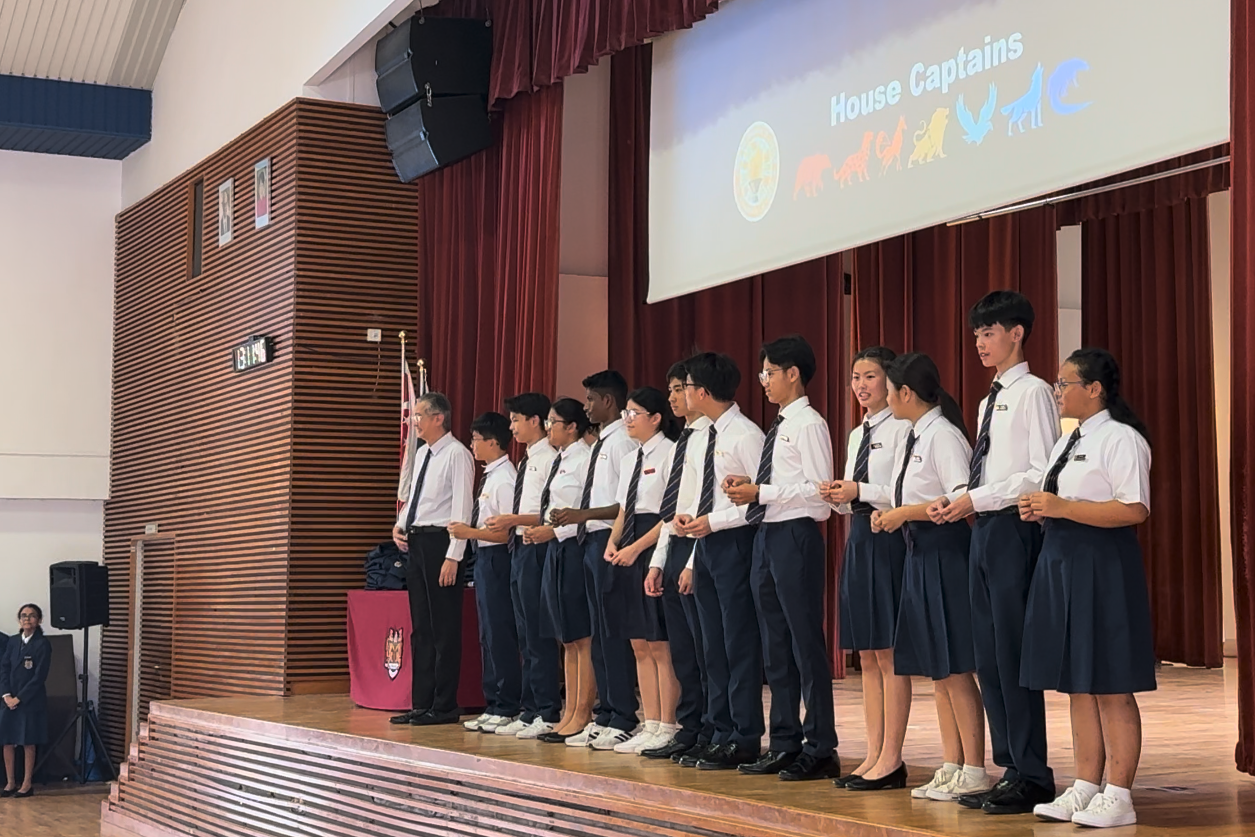Students in white shirts and dark ties stand on a school stage beneath a projected "House Captains" slide.