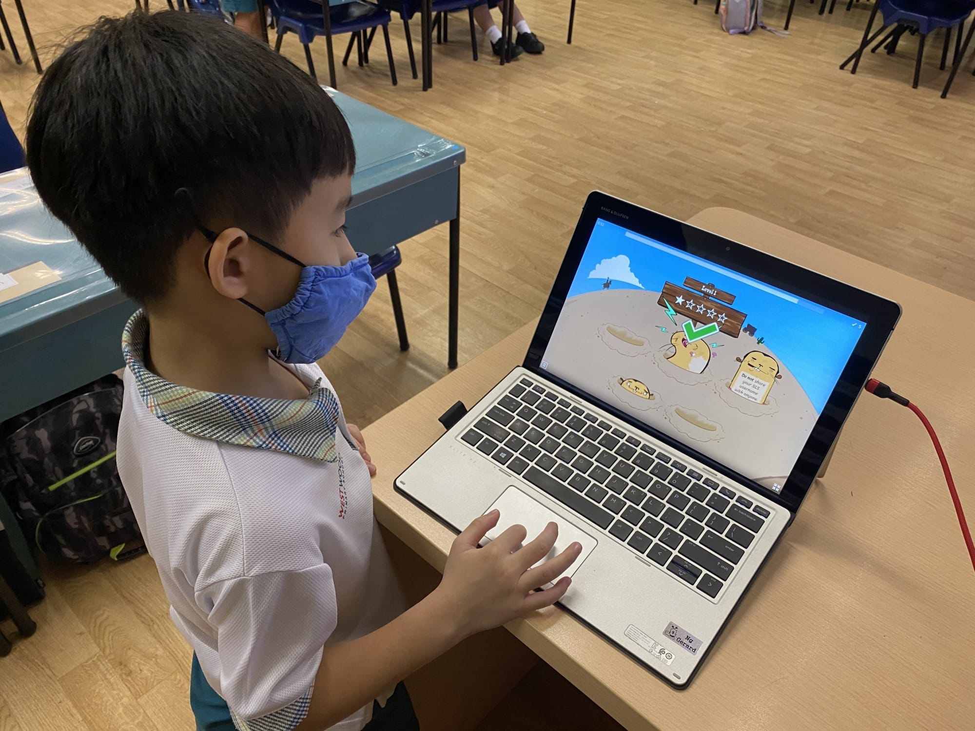 A young boy wearing a face mask uses a laptop displaying a colorful educational game in a classroom.