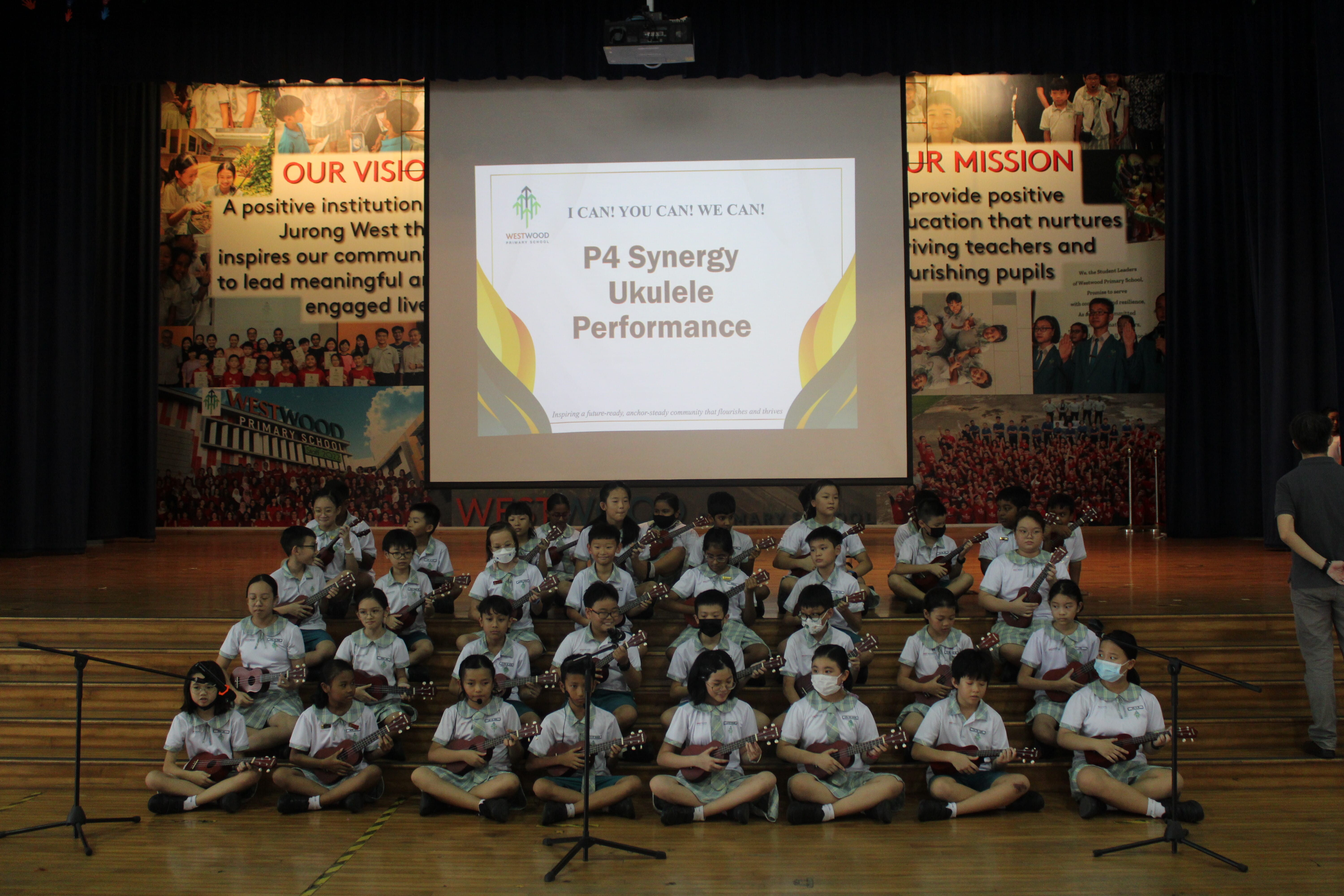Dozens of schoolchildren on stage holding ukuleles, with a screen reading P4 Synergy Ukulele Performance.