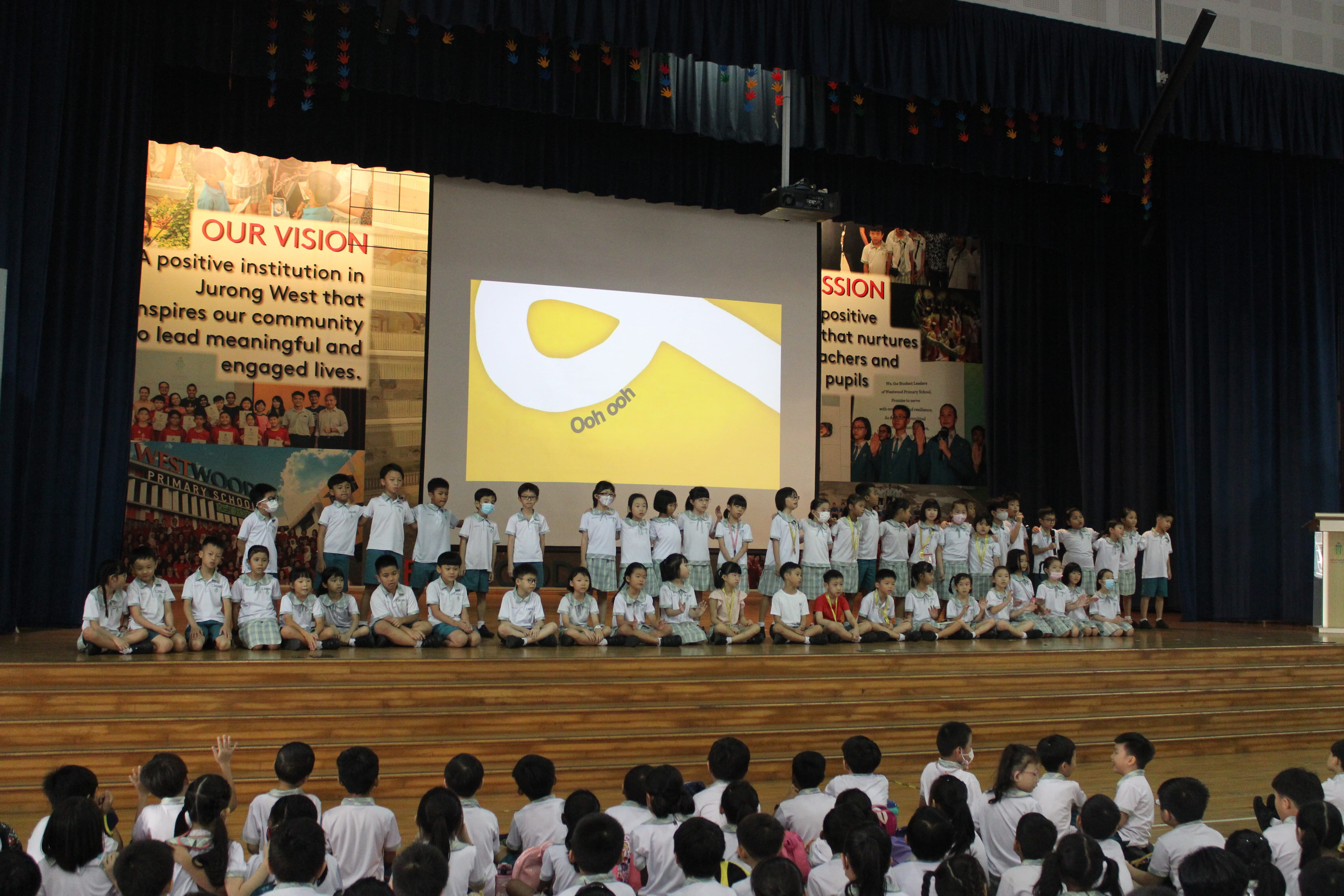 Many schoolchildren in uniforms on a stage under a projector screen, with seated audience in foreground.
