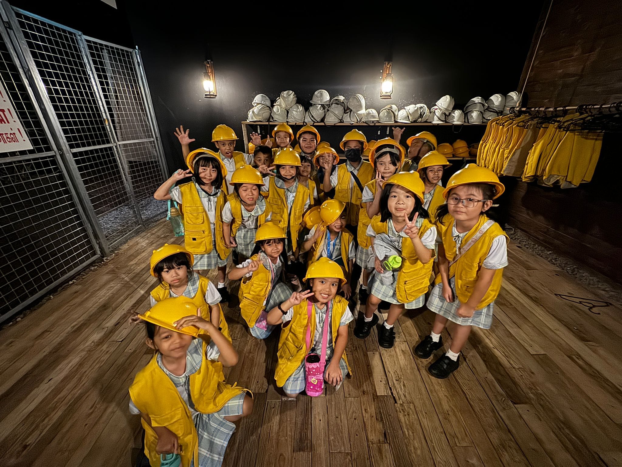 A group of young children wearing yellow hard hats and vests pose and smile inside a dimly lit room.