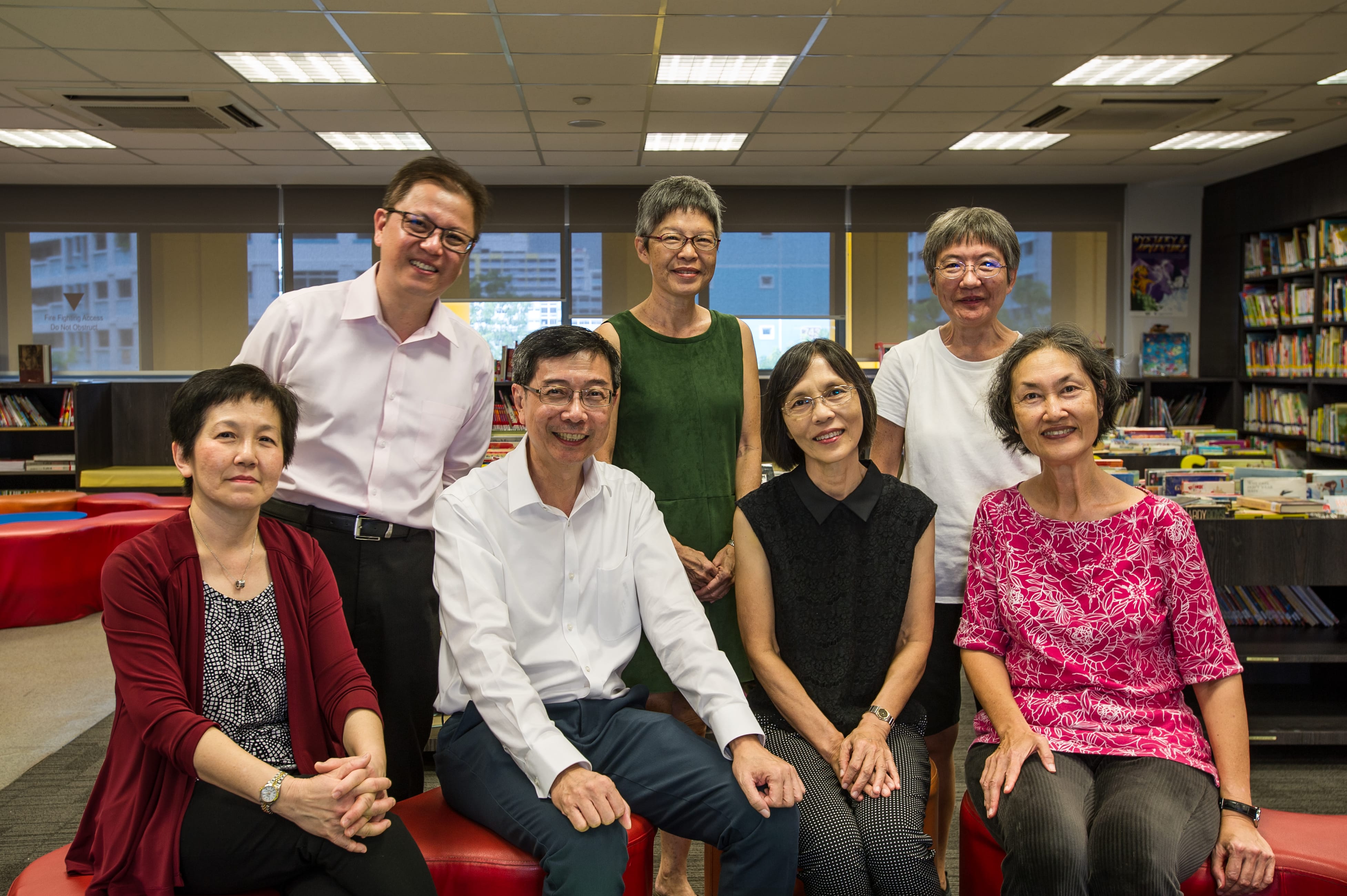 Seven adults pose in a library; four seated on red benches and three standing behind, all smiling.