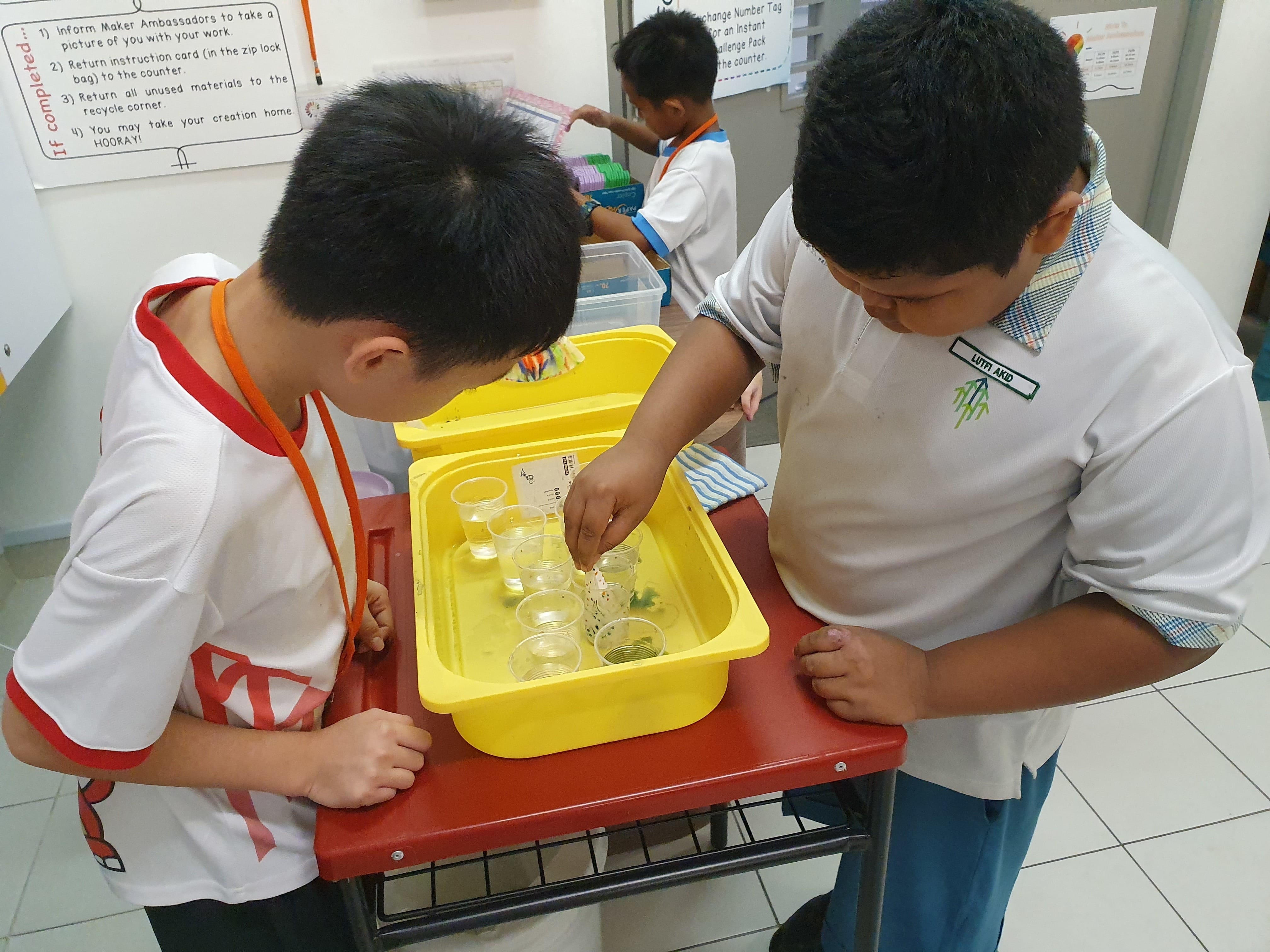 Two school boys examine small plastic cups filled with liquid inside a yellow container on a red cart.