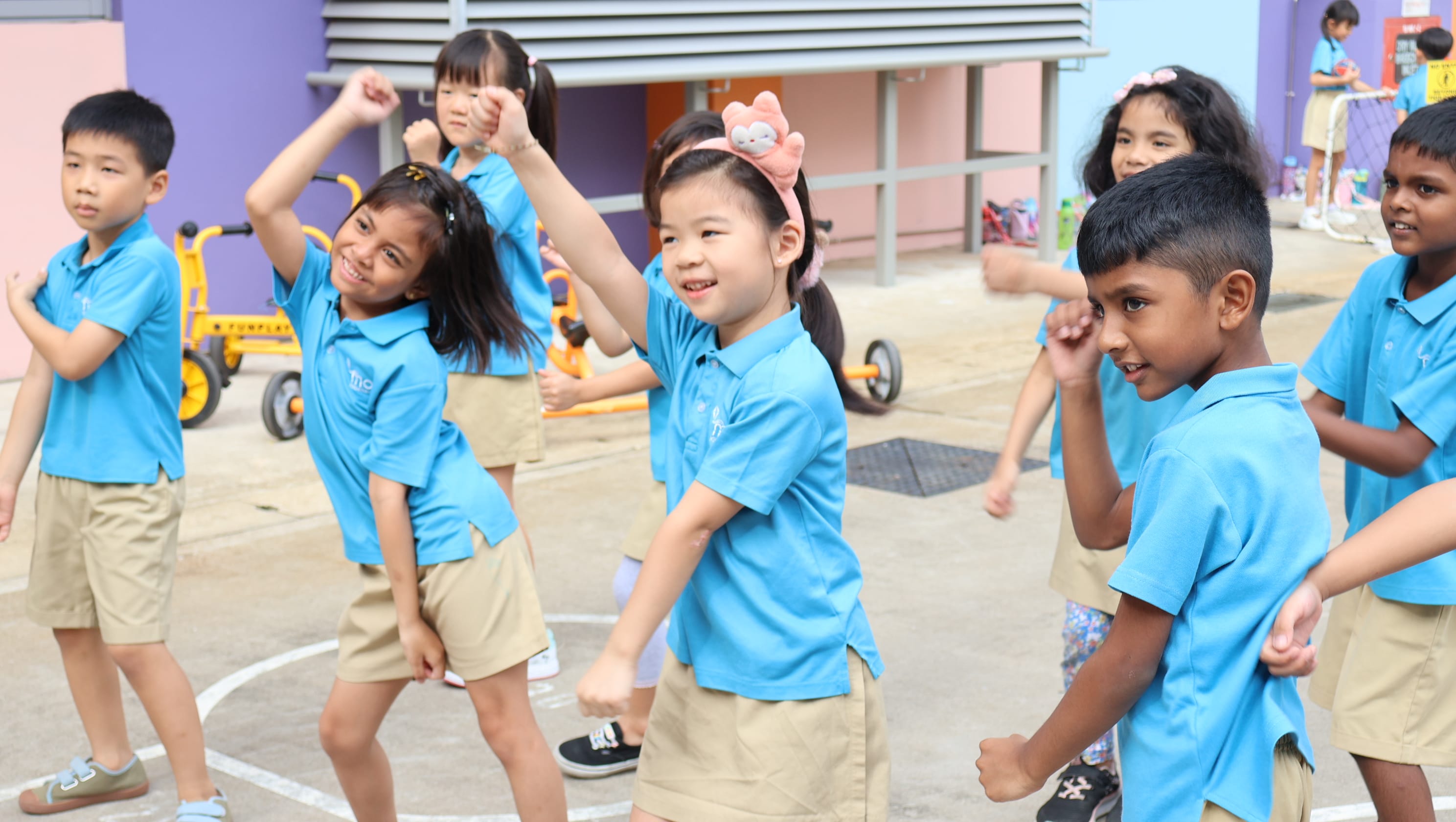 Several young children in blue polo shirts and khaki shorts dance together outdoors in a school playground.