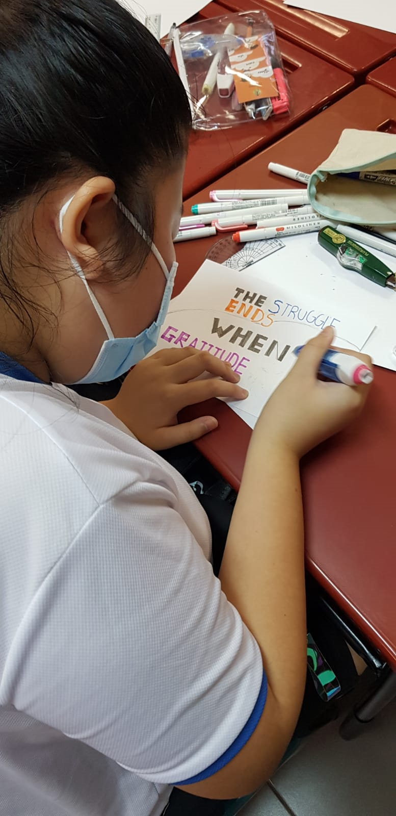 A masked child writes the phrase "The Struggle Ends When Gratitude" on paper using colorful markers at a desk.