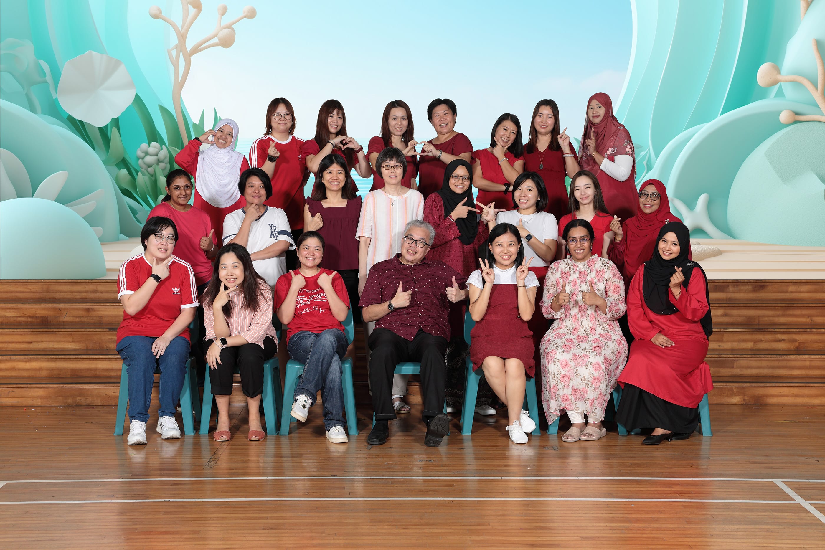 A large group of about 25 people posing in rows, mostly wearing red, smiling and making hand gestures indoors.