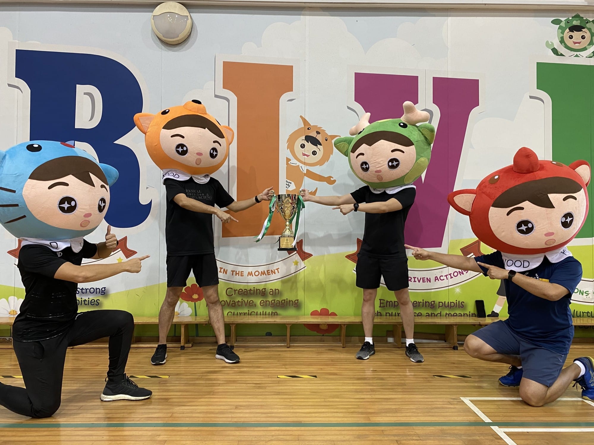 Four people wearing large cartoon character heads pose with a trophy in a school gymnasium.