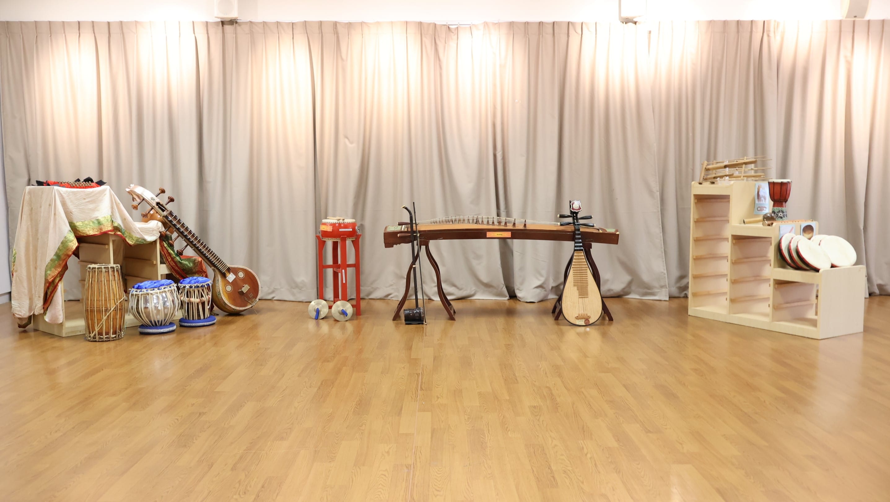 Various traditional Asian instruments including a guzheng, pipa, sitar, and tabla drums displayed in a room with beige curtains.