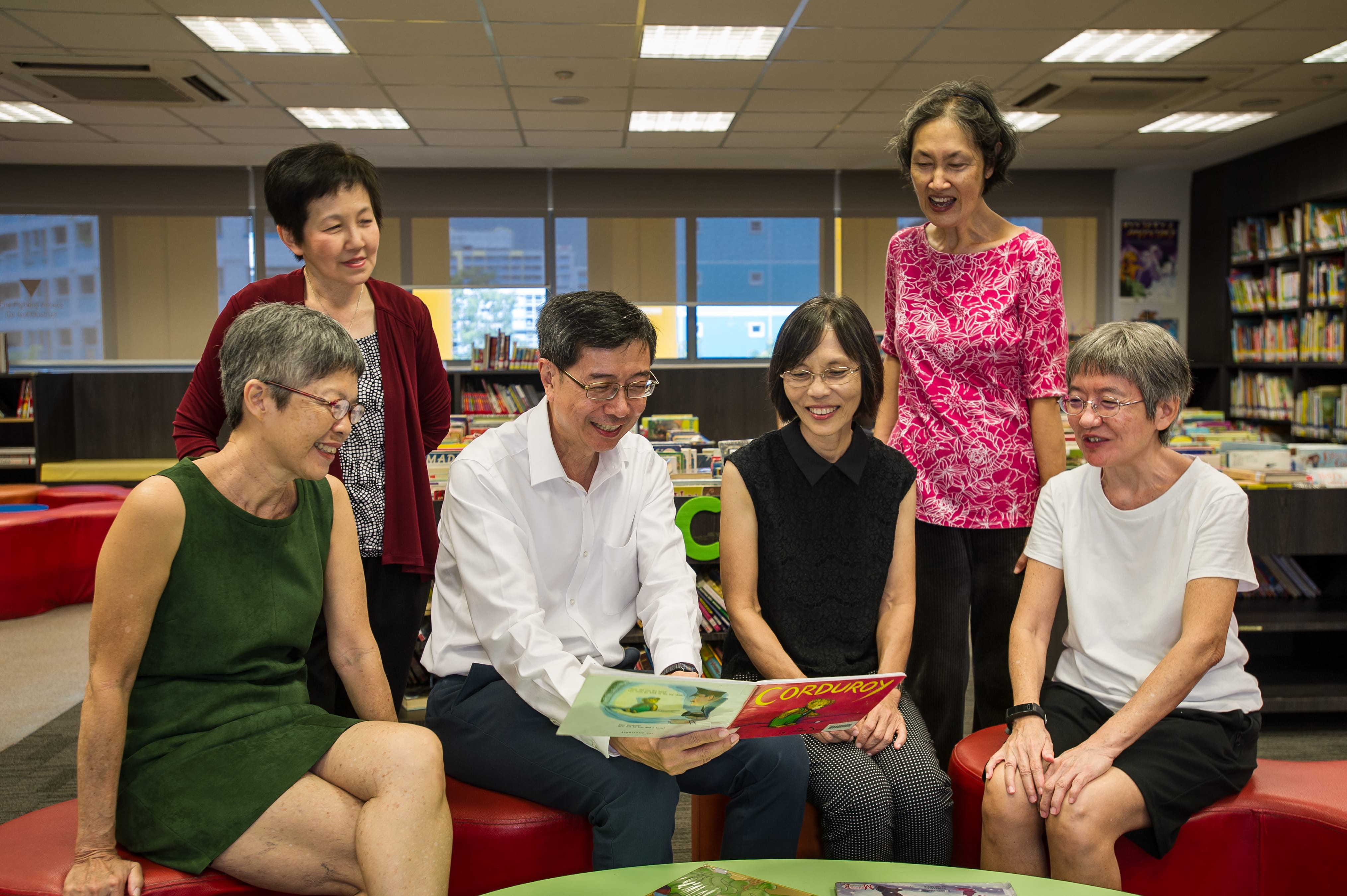 Five adults sit and stand in a library, smiling as a man reads a picture book to the group.