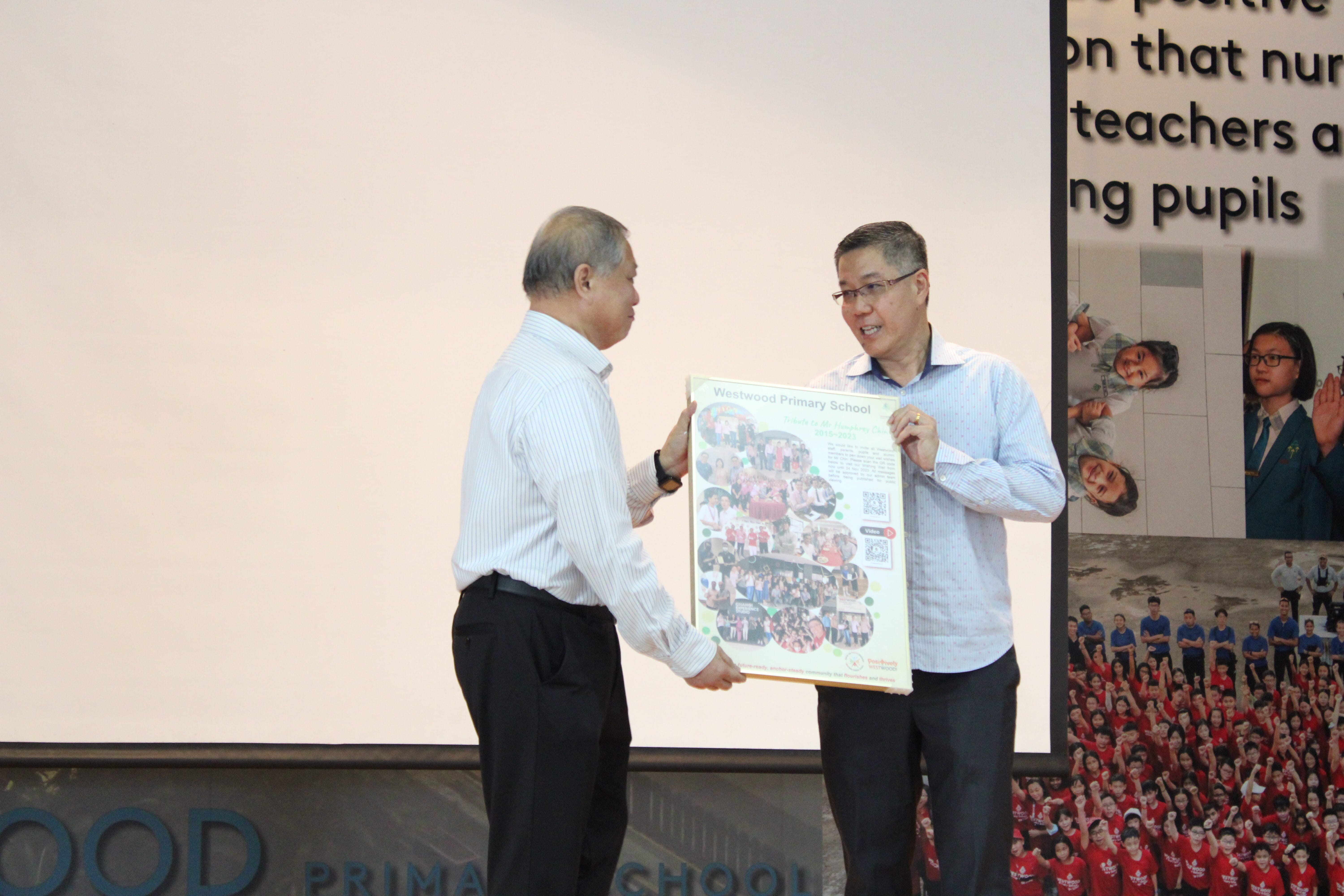 Two men on a stage hold a Westwood Primary School framed photo collage together during a presentation.
