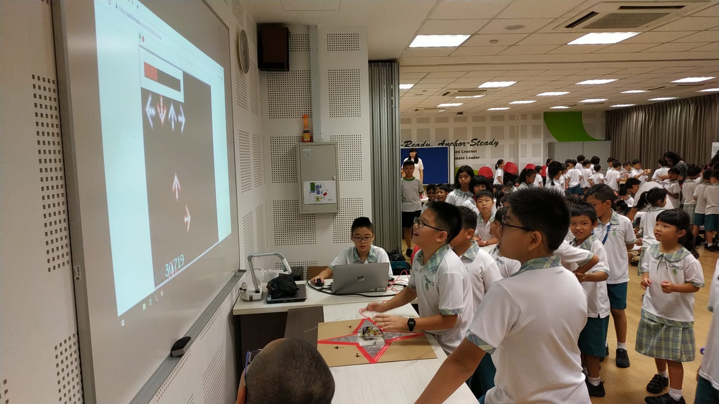 Students in school uniforms gather around a laptop and projected screen showing arrows during a classroom activity.