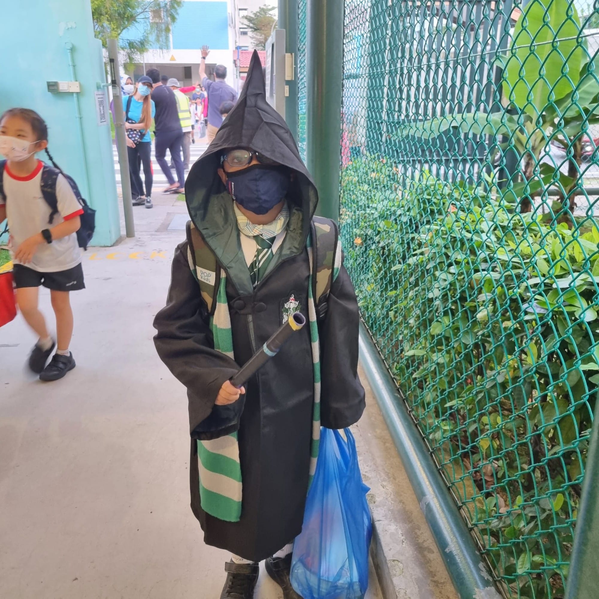 Child in a black wizard robe, pointed hood, green scarf, and mask holds a wand near a chain-link fence.