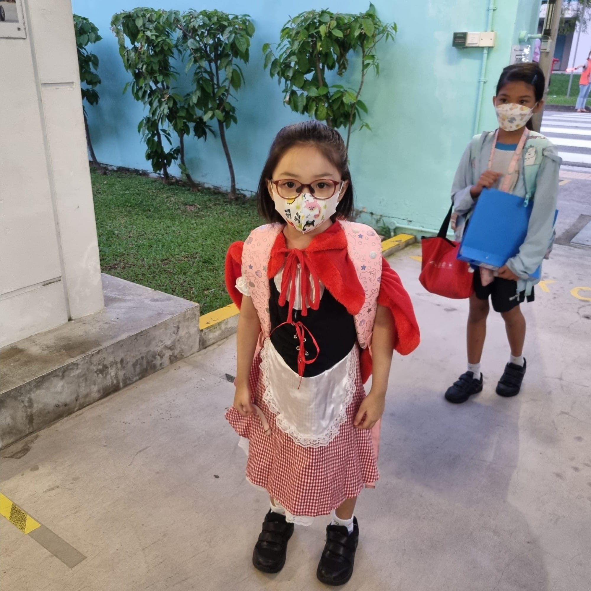 Two children wearing face masks stand outdoors, one dressed in a Red Riding Hood costume with a red cape and plaid skirt.