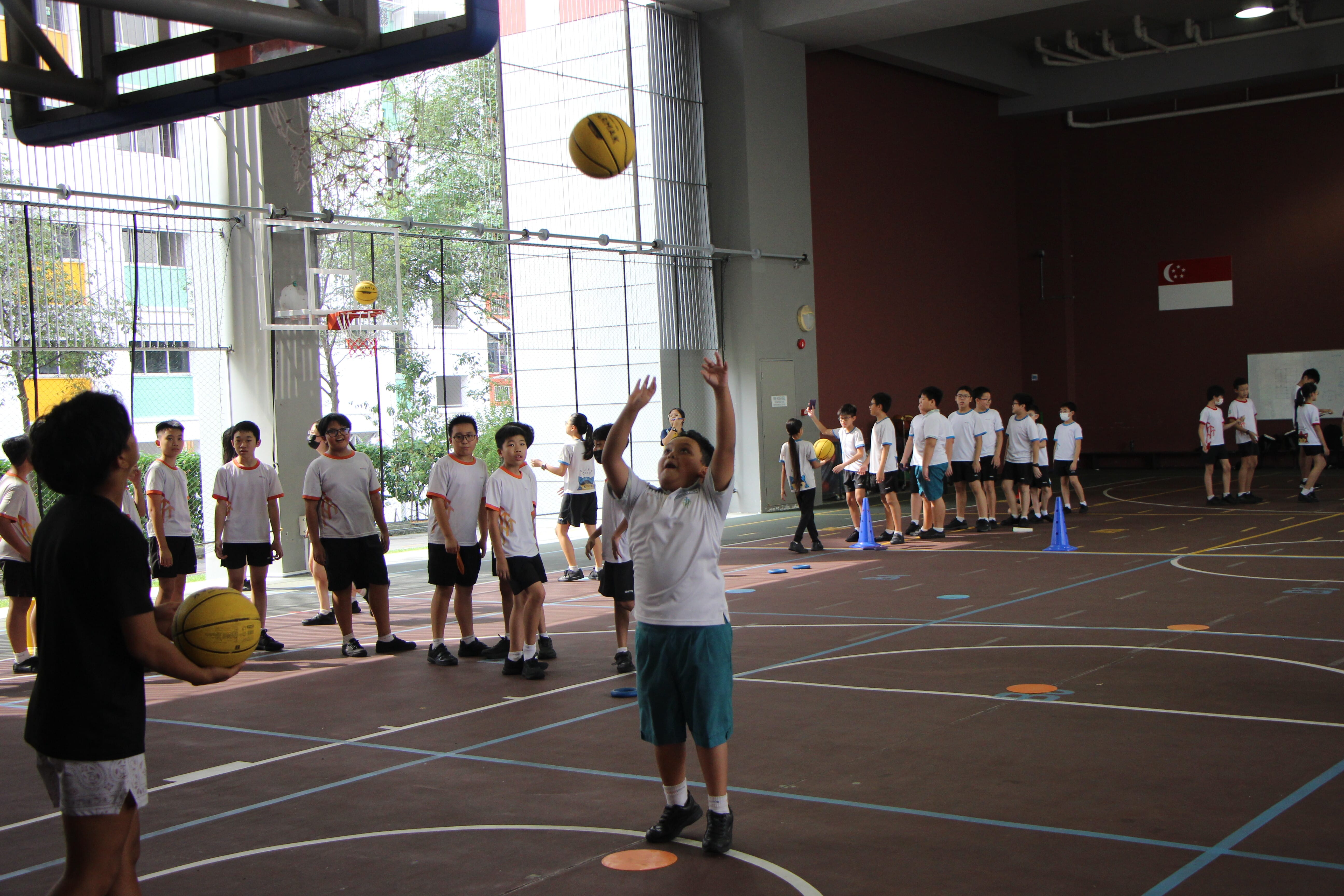 A student in white shirt shoots a basketball during a school PE class indoors with many classmates watching.