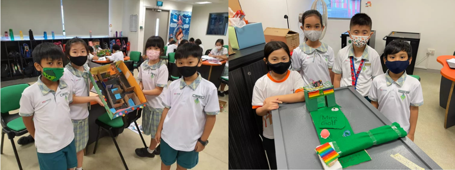 Six masked young students in school uniforms display handmade cardboard projects, including a mini golf course, in a classroom.