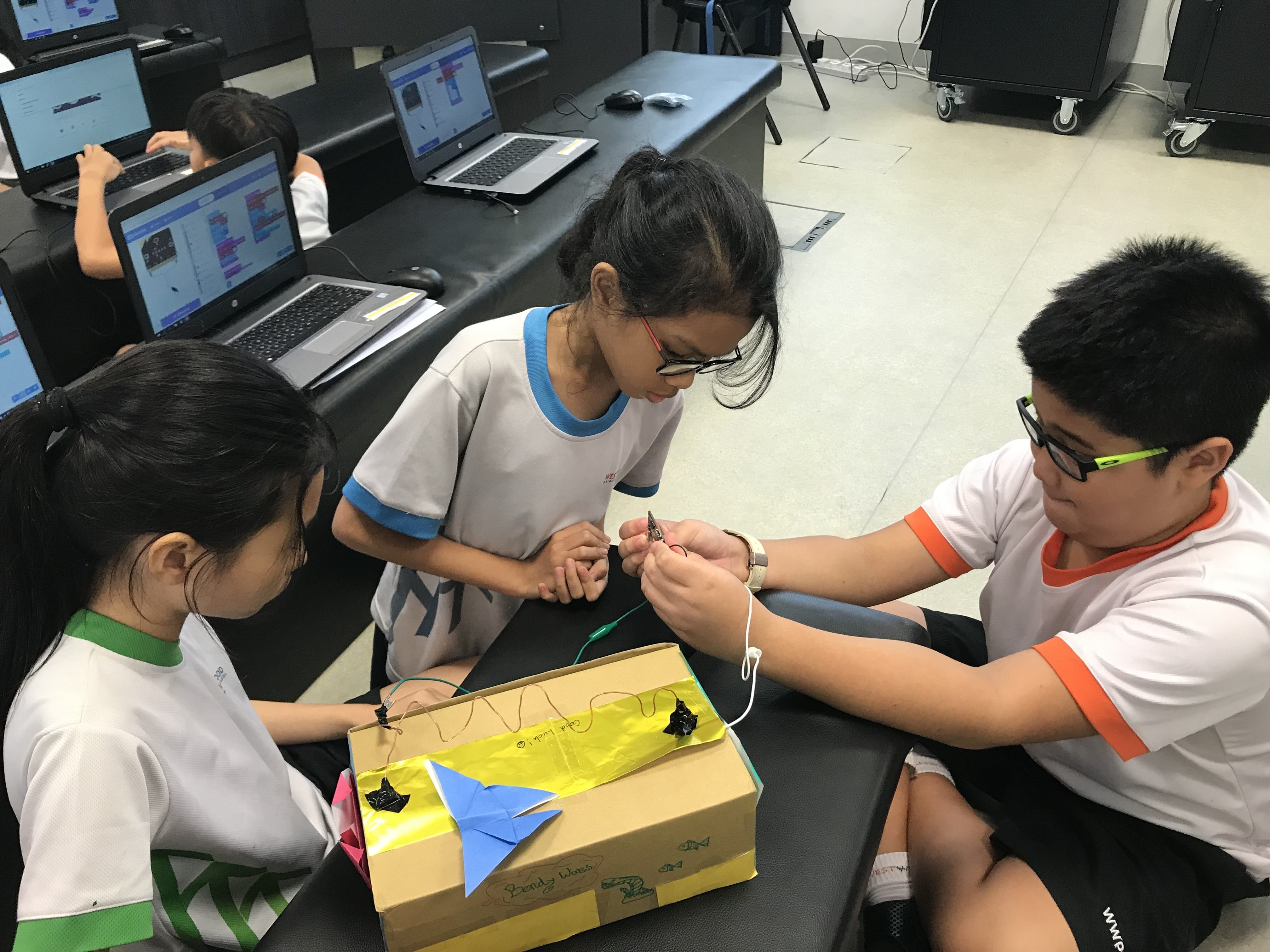 Three children in school uniforms work together on a cardboard box project with origami decorations in a computer lab.