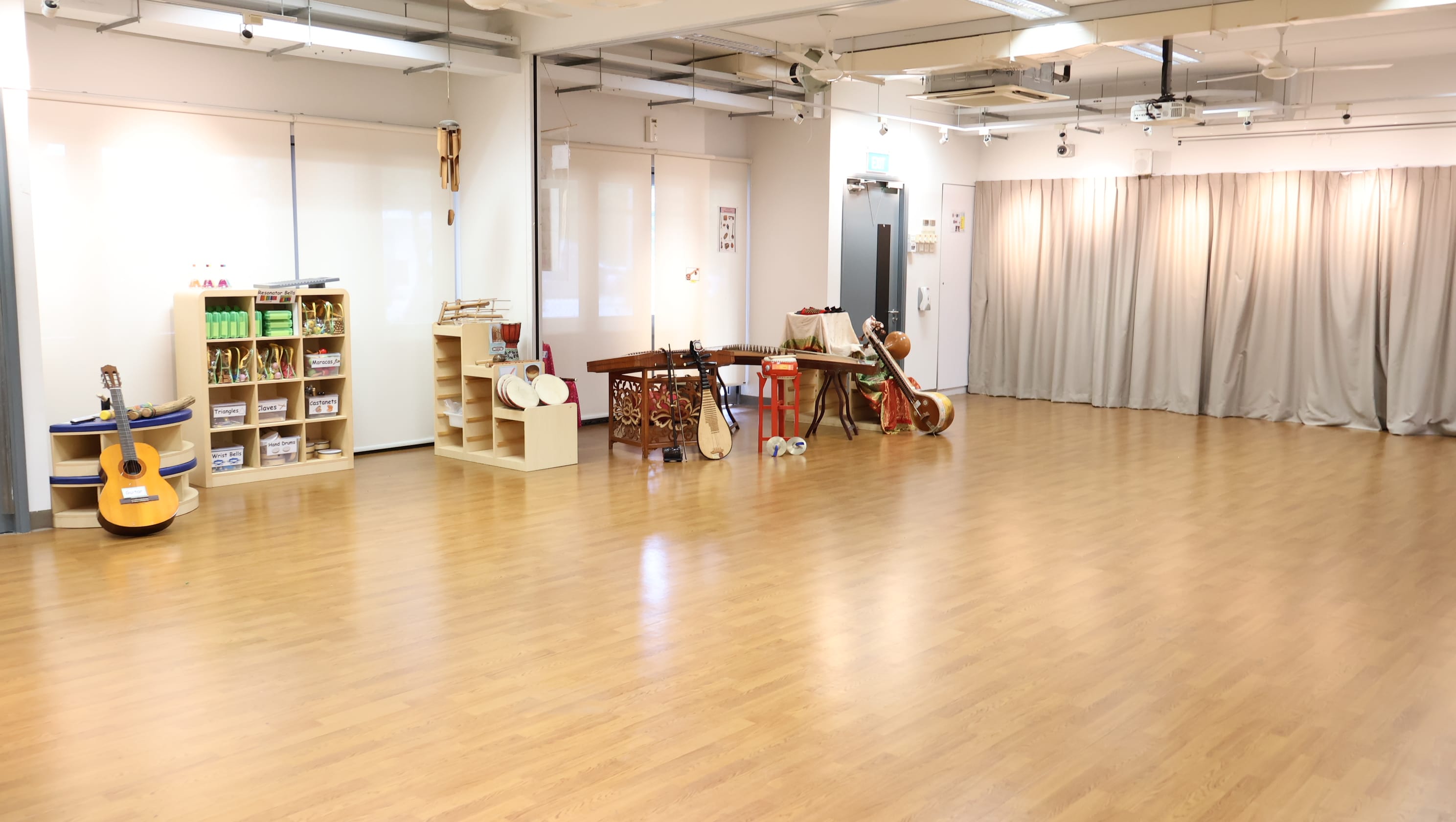 Spacious music room with wood floor, shelves of instruments, a guitar, string instruments, and gray curtains along the wall.
